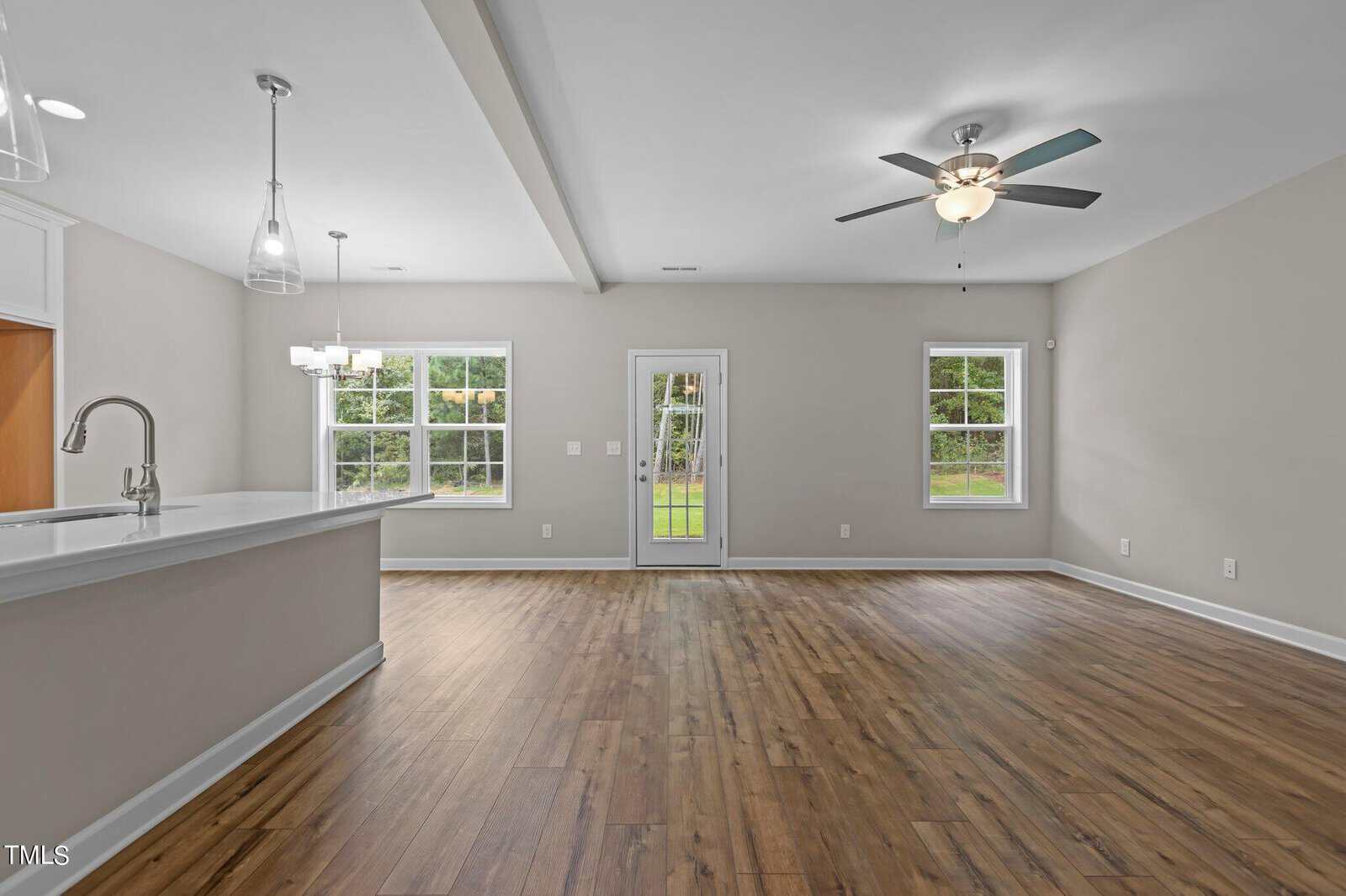 Open-concept living and dining area with light wood floors, neutral walls, two windows, a glass door to the backyard, ceiling fan, and modern pendant lights above a white kitchen counter.