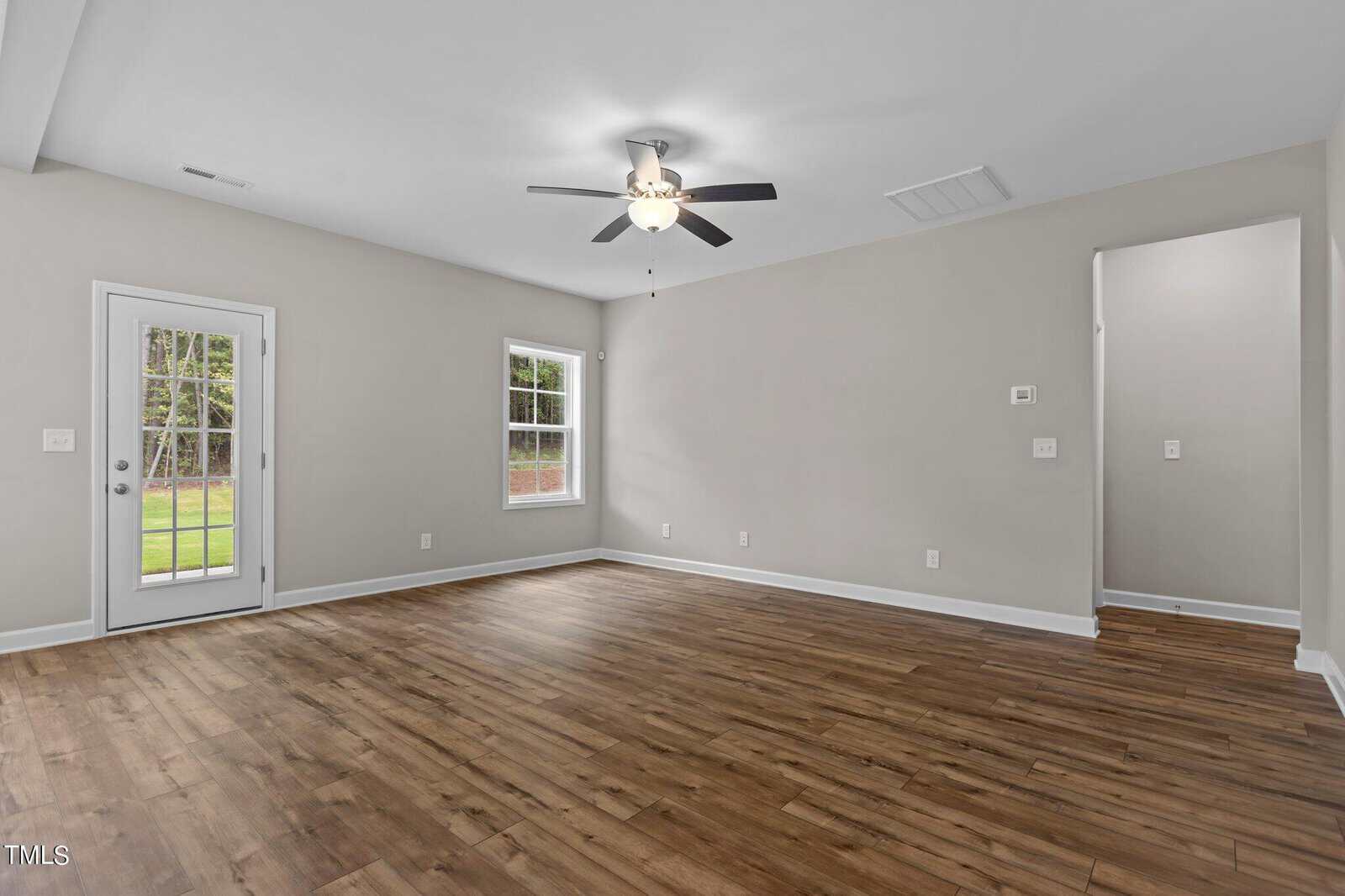 Empty room with light brown wood flooring, beige walls, a ceiling fan with lights, a window, a glass door leading outside, and an open doorway to another room. Natural light enters through the window and door.