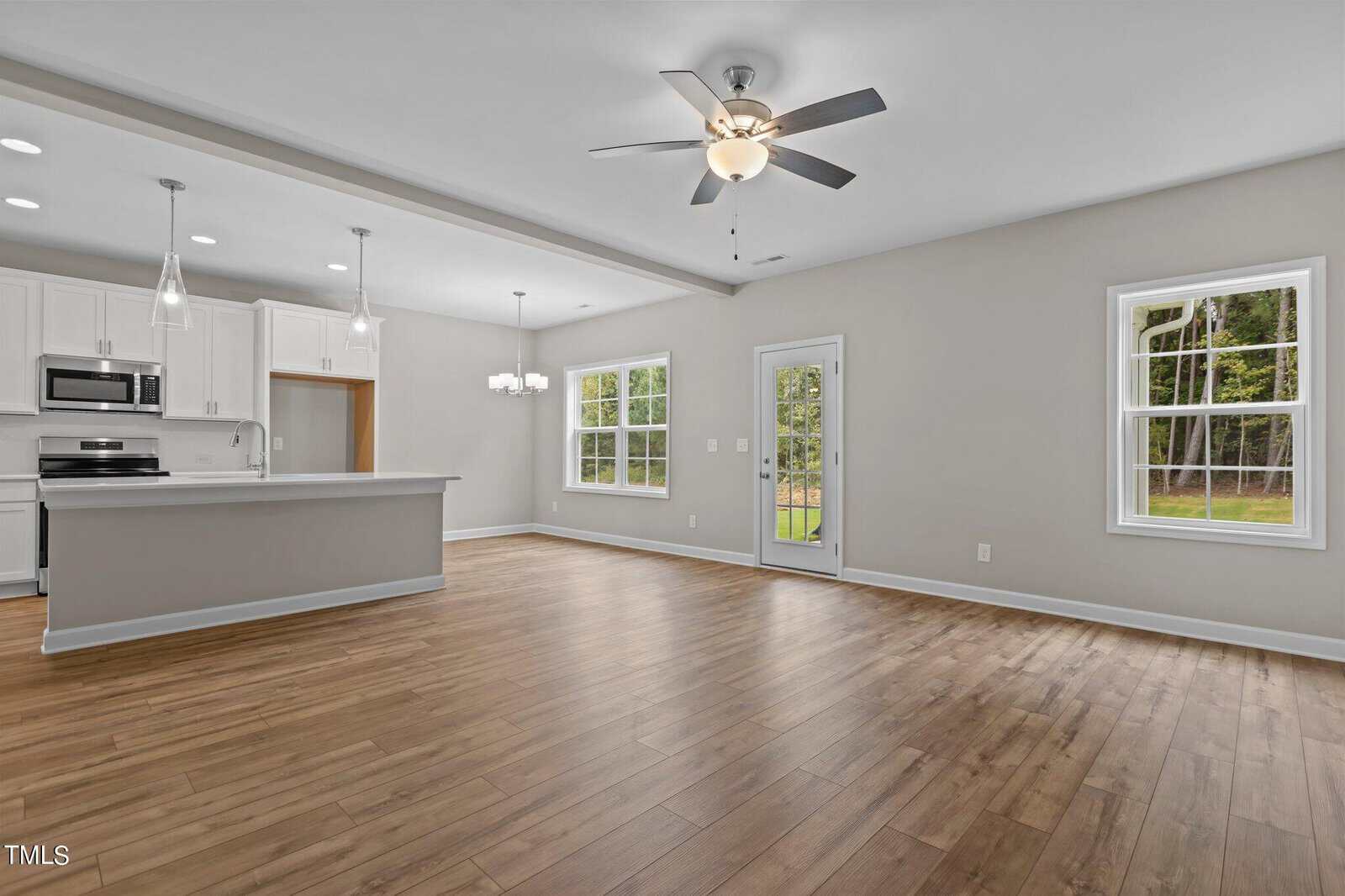 Open-concept kitchen and living area with wood flooring, white cabinets, stainless steel appliances, ceiling fan, large windows, and a glass door leading to a backyard with trees outside. Room is empty and well-lit.