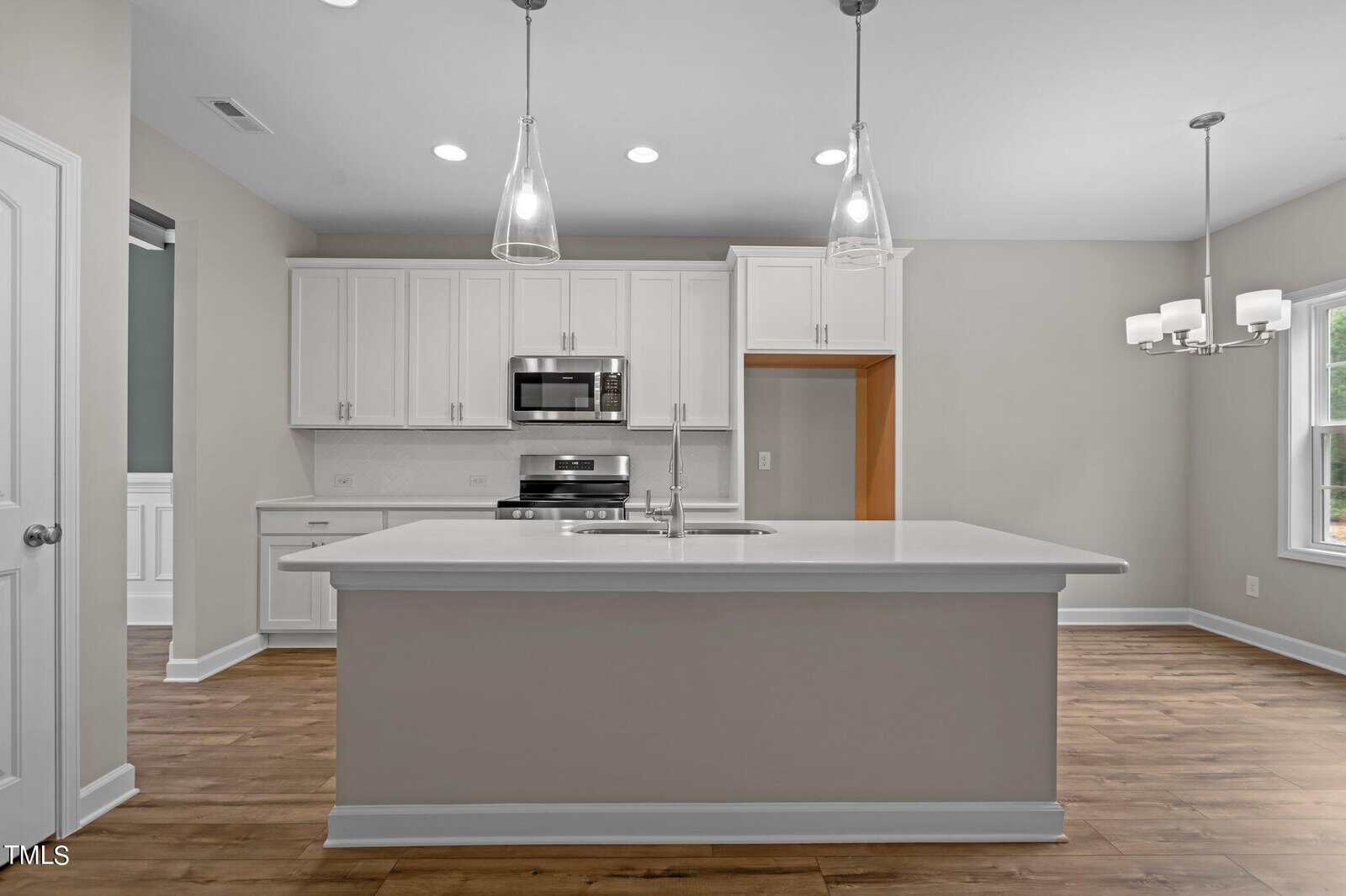 Modern kitchen with white cabinets, stainless steel appliances, a large island with sink, pendant lights above, wood flooring, and an adjacent dining area with a window and a chandelier.