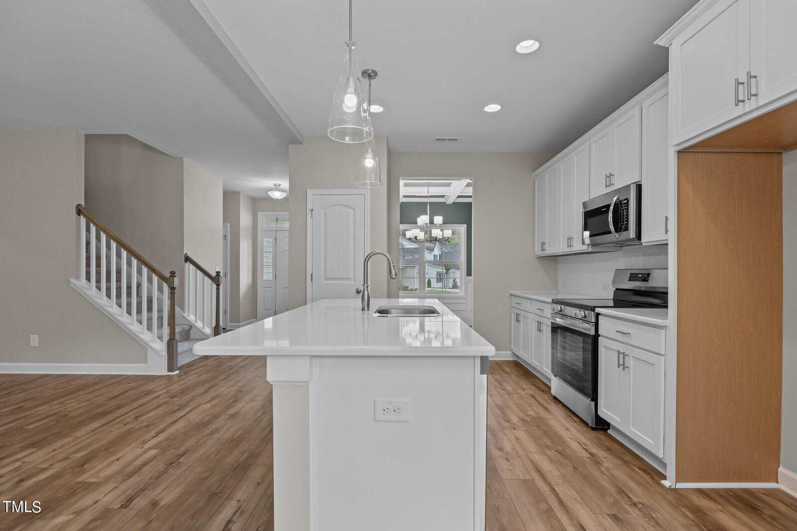 Modern kitchen with white cabinets, stainless steel appliances, a center island with sink, pendant lights, and wood flooring. The kitchen opens to a staircase and a hallway, with neutral-colored walls and ample natural light.