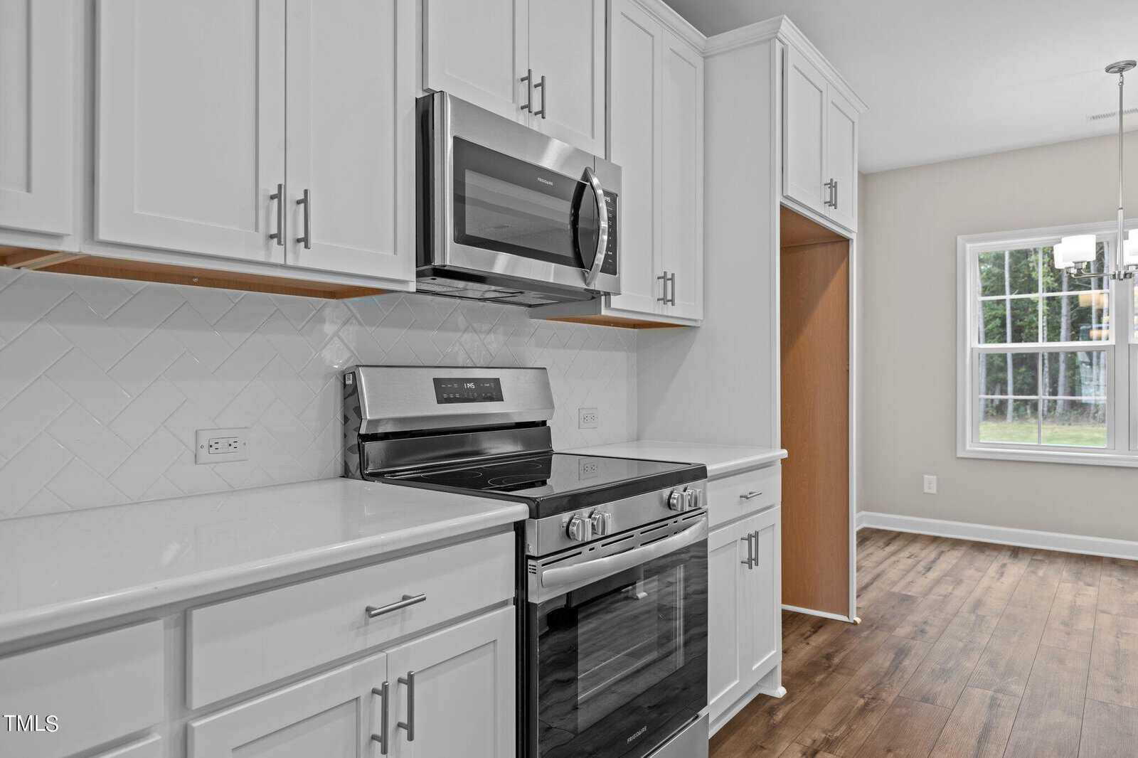 Modern kitchen with white cabinets, stainless steel appliances including a microwave and stove, white tile backsplash in a herringbone pattern, and wood flooring. A window lets in natural light.