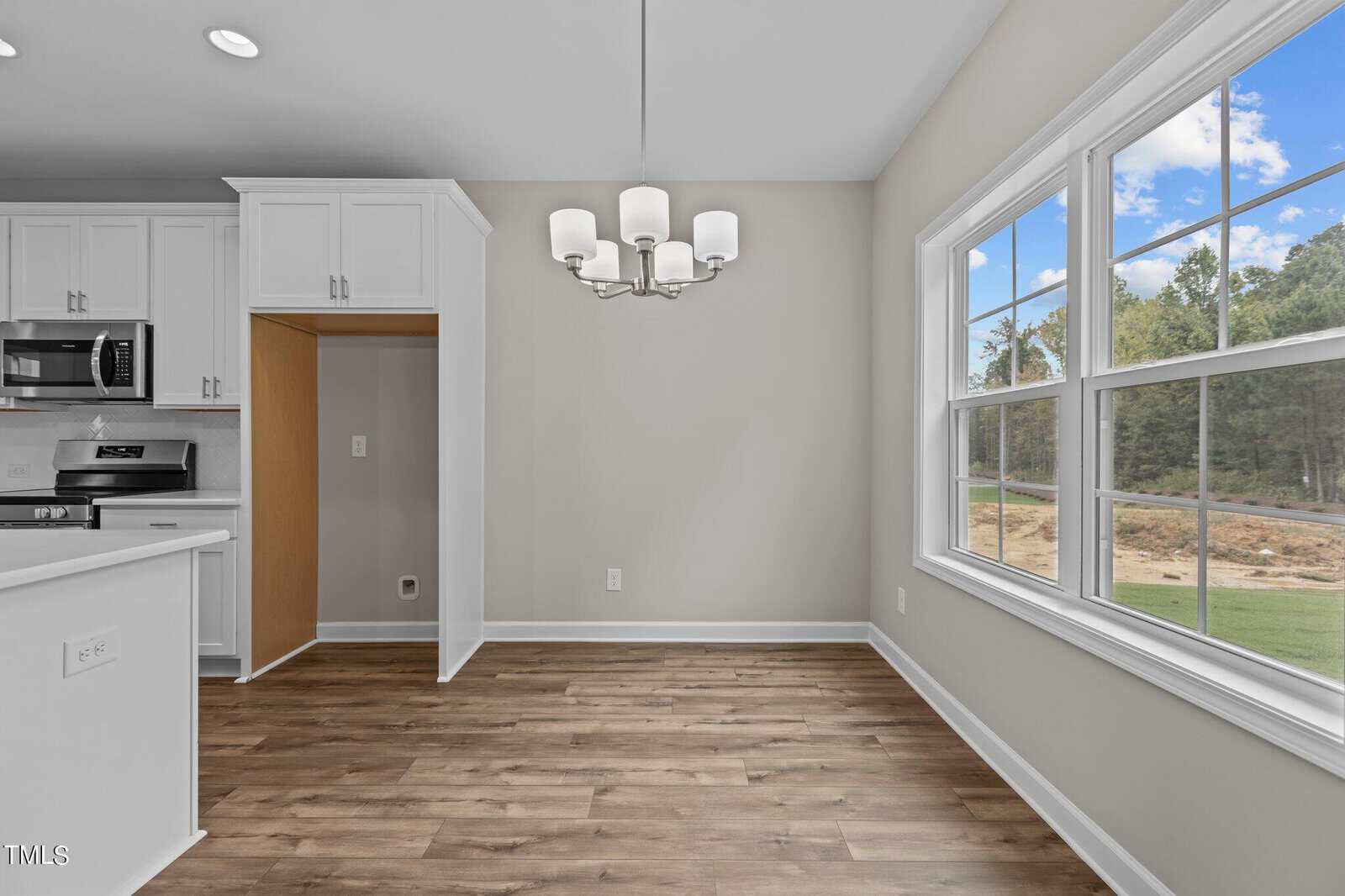 A bright, empty dining area with wood floors, a modern chandelier, large windows showing trees outside, and an adjacent kitchen with white cabinets and stainless steel appliances.