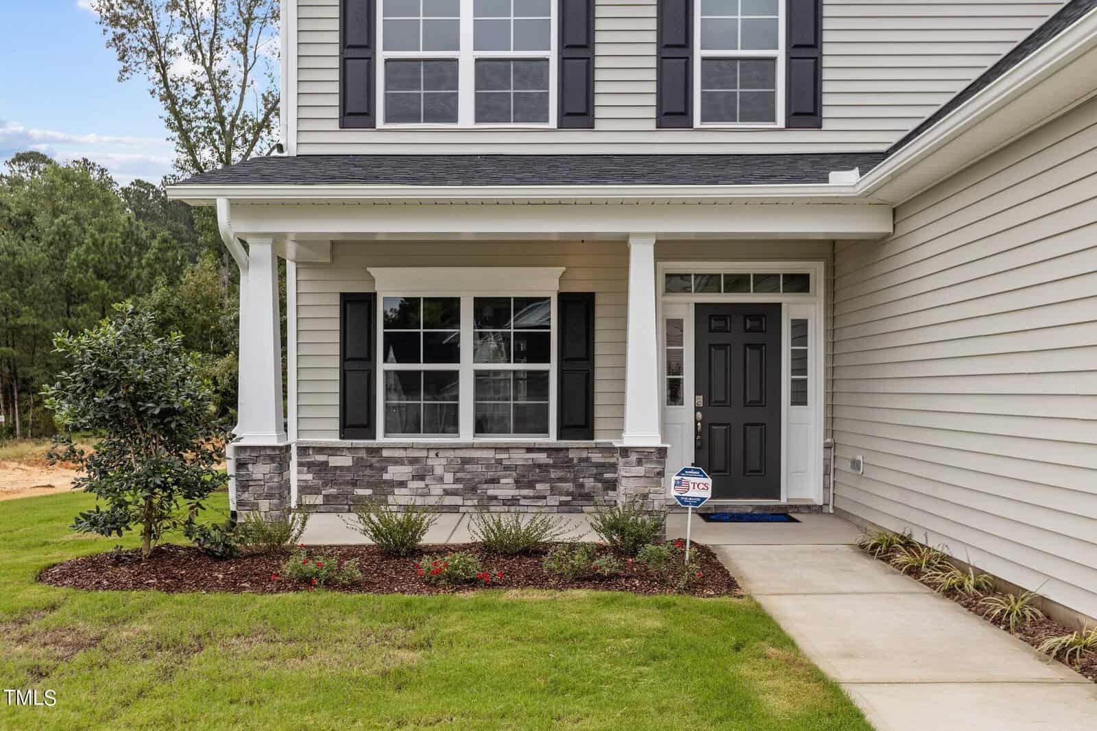 A house with gray siding, black shutters, a covered front porch, decorative stonework, a black front door with glass panels, small shrubs, a manicured lawn, and a home security sign by the entrance.