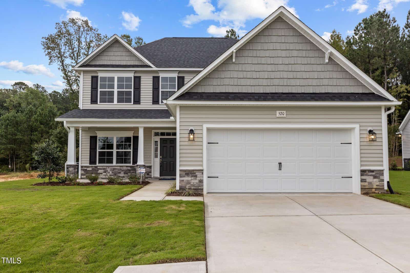 Two-story suburban house with gray siding, white trim, a covered front porch, large double garage, and a well-manicured lawn, set against a backdrop of trees and a partly cloudy sky.