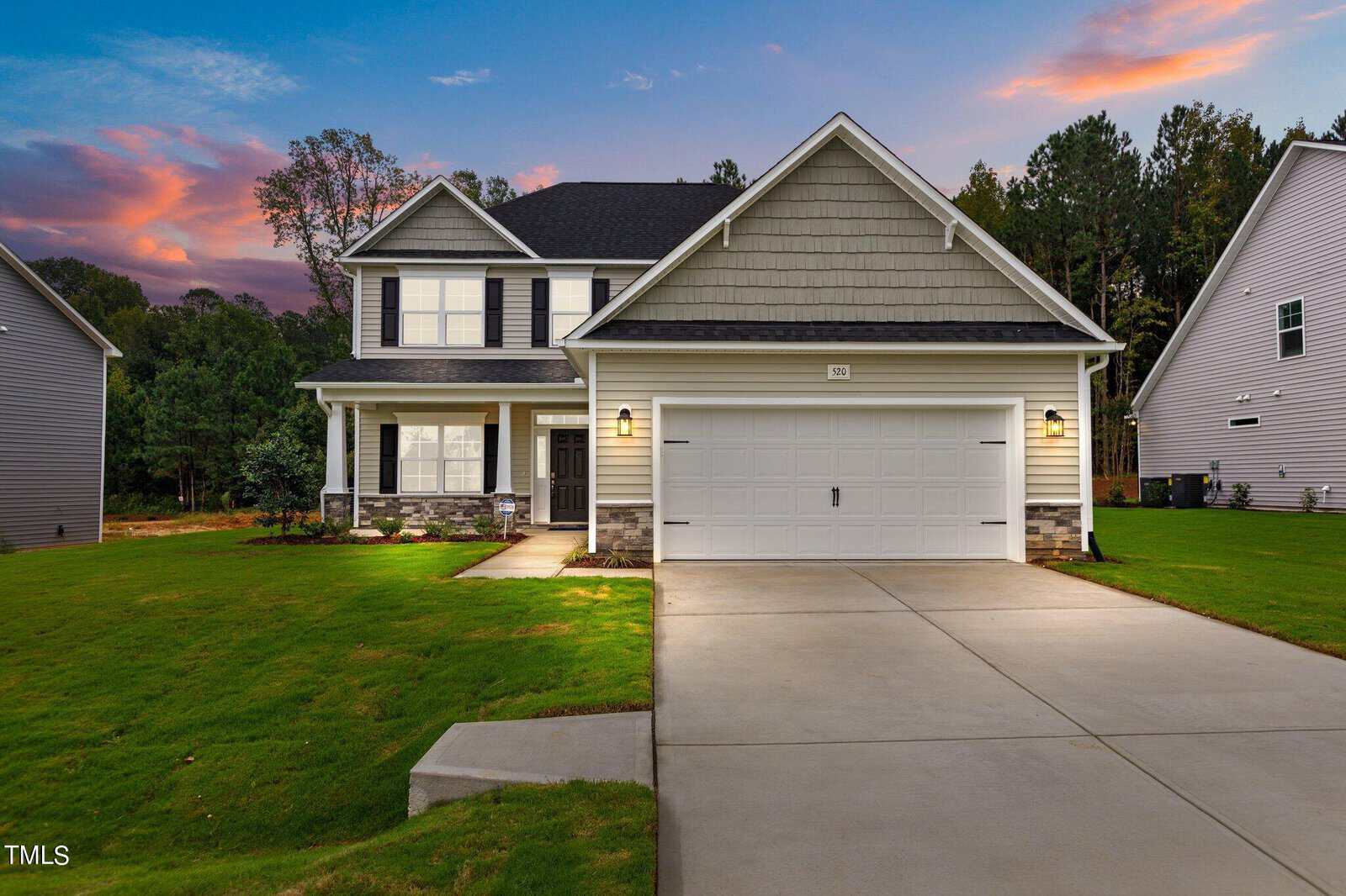 A modern two-story house with a large front lawn, double garage, porch, and exterior lights on at dusk. The sky has orange and purple hues, and trees surround the property.