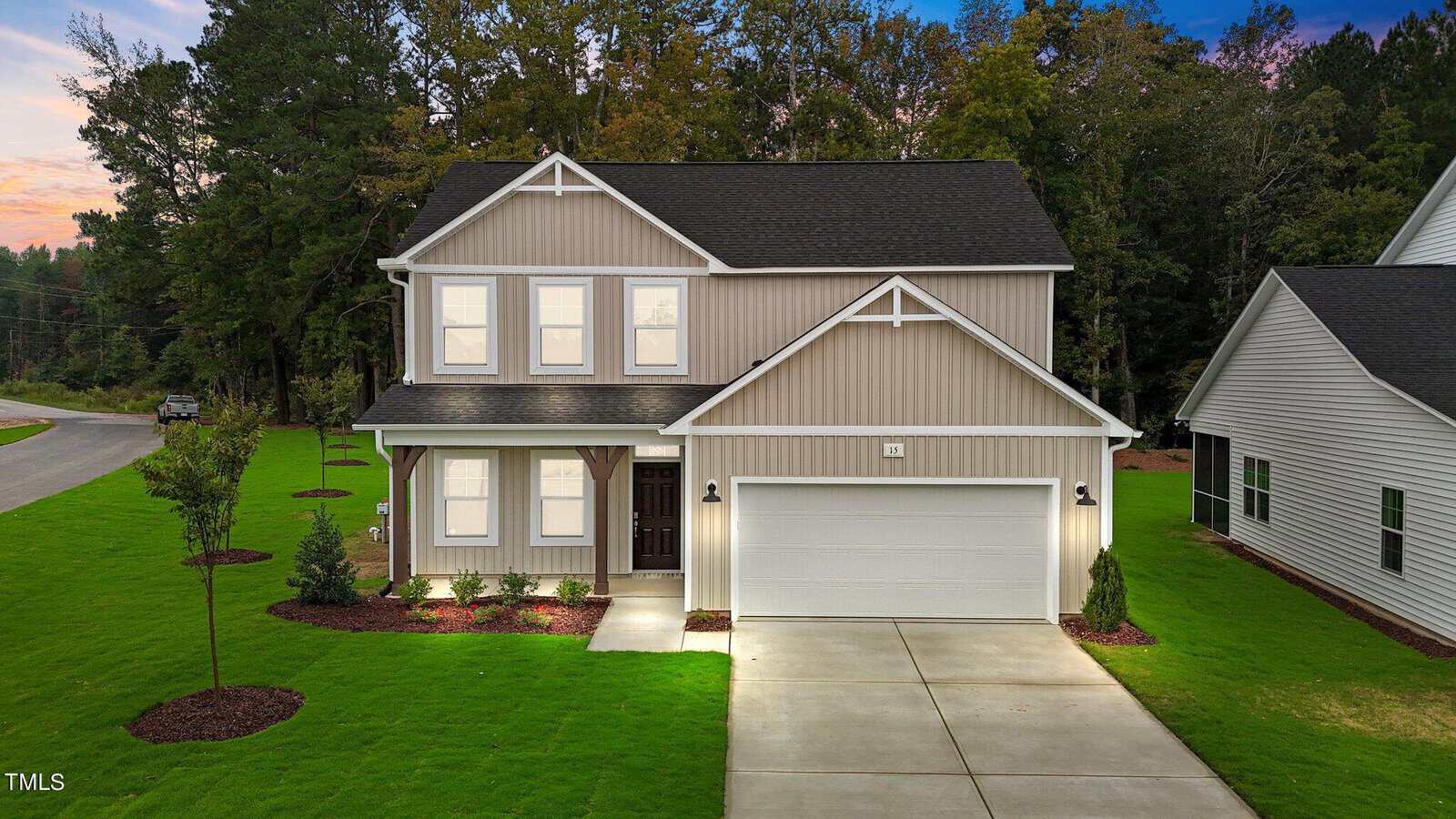 Two-story beige house with white trim, attached two-car garage, front porch, and well-maintained lawn with young trees and shrubs, set against a backdrop of tall trees at dusk.