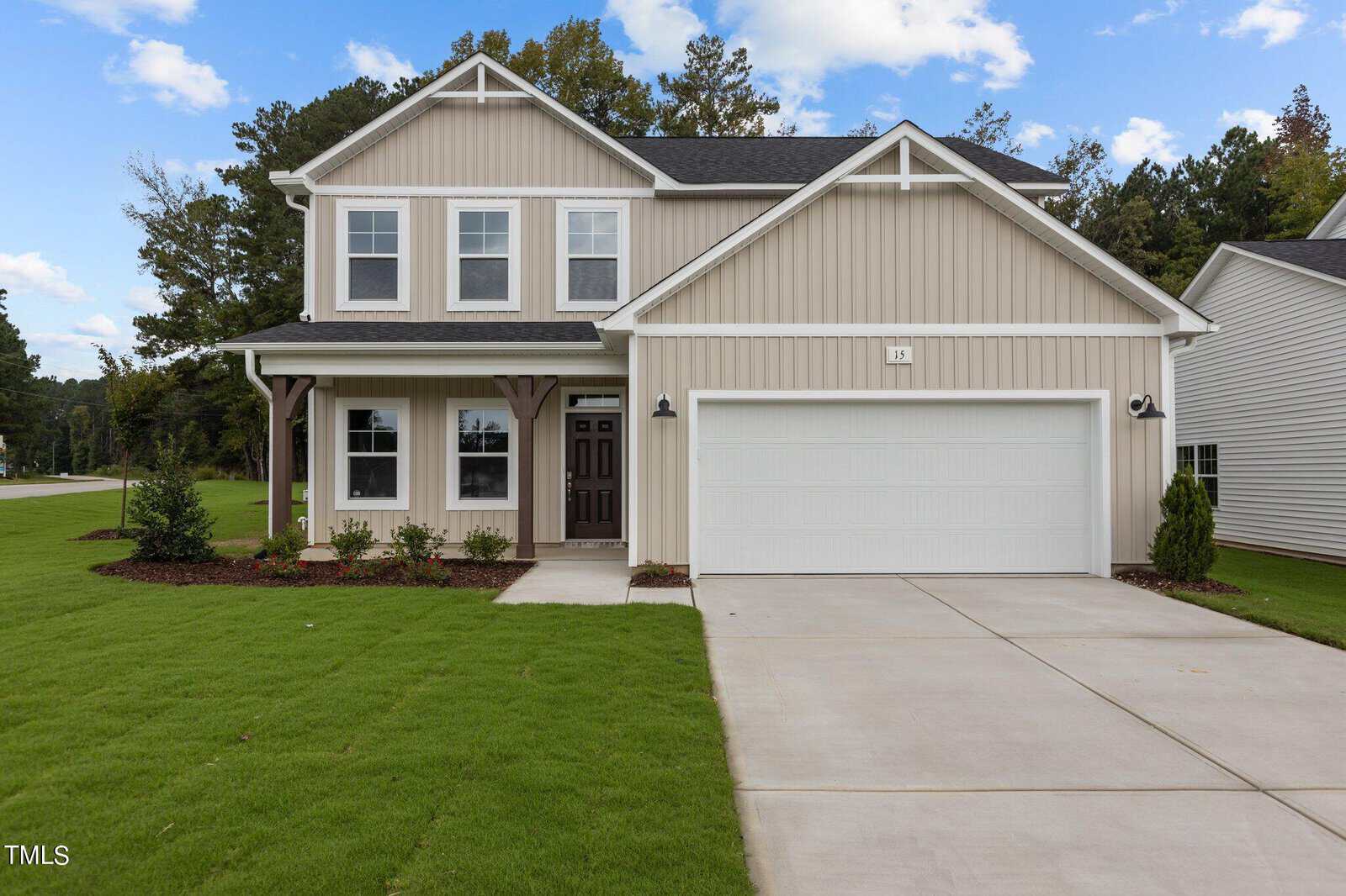 Two-story beige house with white trim, covered front porch, double garage, and neatly landscaped yard with green grass and shrubs, set against a backdrop of trees and blue sky with clouds.