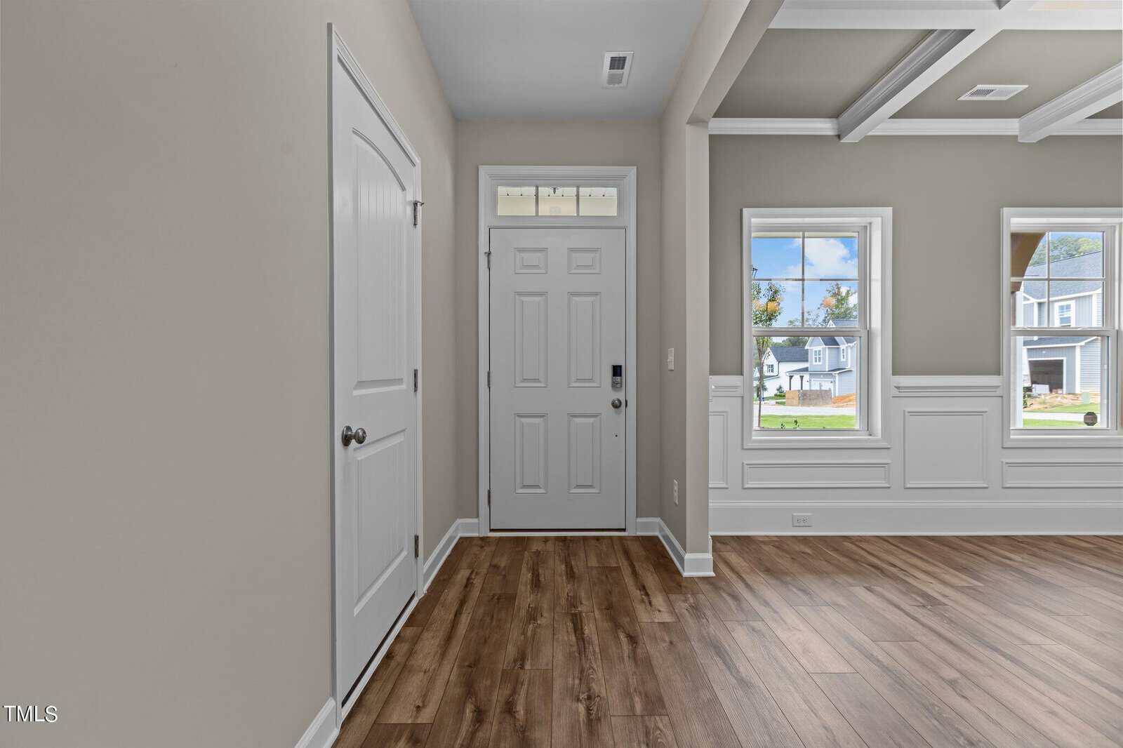 A modern entryway with a white front door, sidelights, a closet door, beige walls, wood flooring, and large windows letting in natural light, offering a view of houses outside.