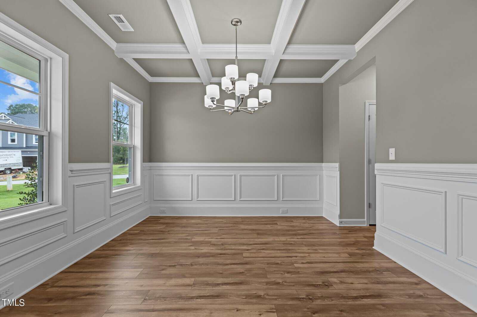 A bright, empty dining room with gray walls, white wainscoting, coffered ceiling, wood floor, windows letting in natural light, and a modern chandelier hanging in the center.