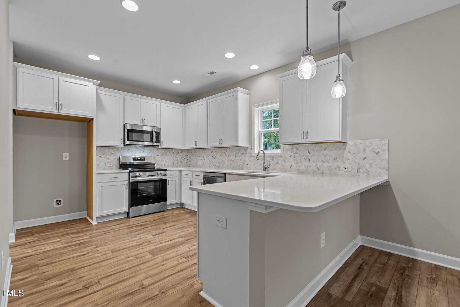 Modern kitchen with white cabinets, stainless steel appliances, marble-patterned backsplash, wood flooring, and two pendant lights over a large white peninsula countertop. A window provides natural light above the sink.