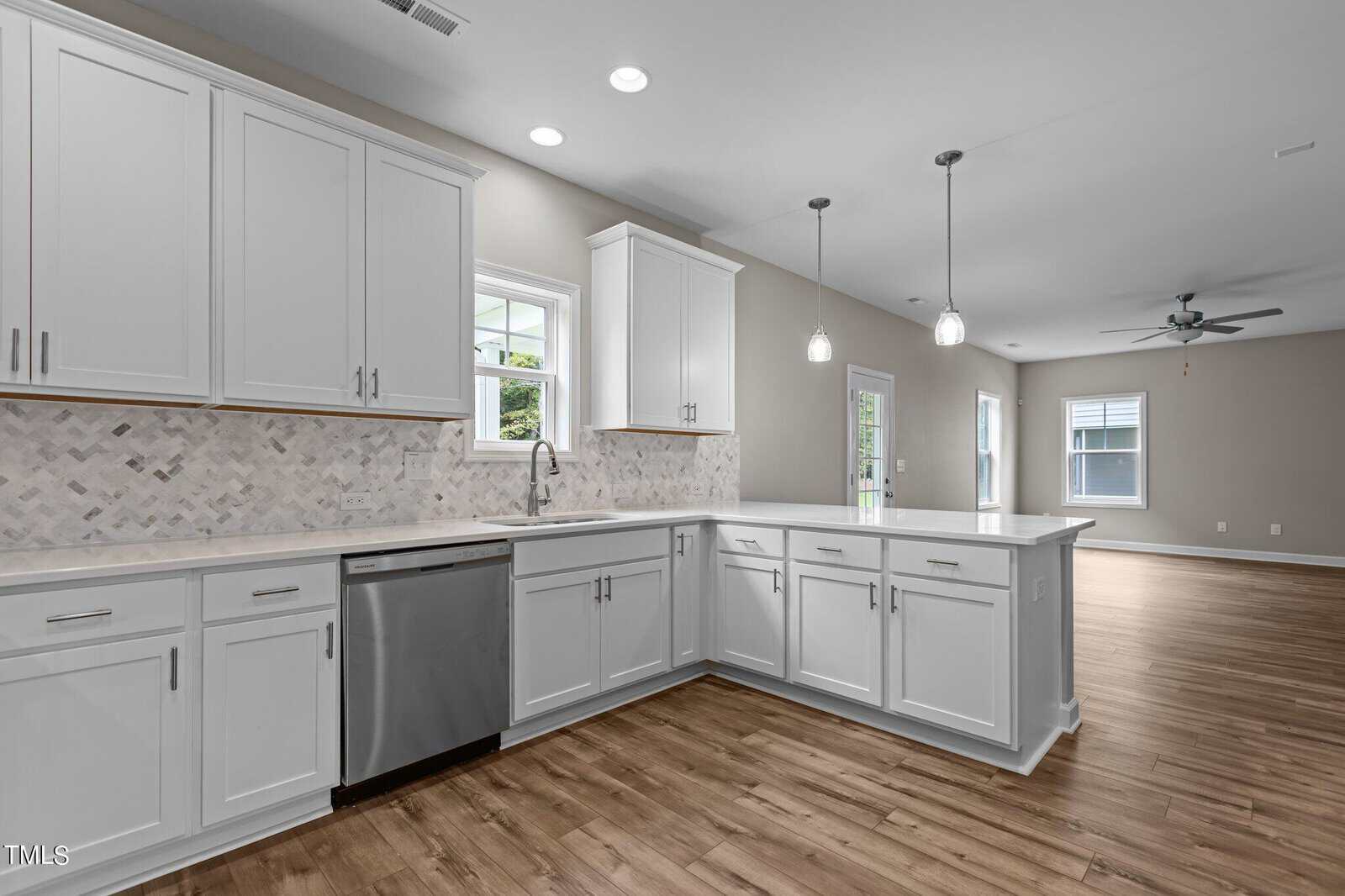 Modern kitchen with white cabinets, stainless steel dishwasher, tile backsplash, and light wood flooring, open to a spacious living area with large windows, neutral walls, and pendant lighting over the counter.