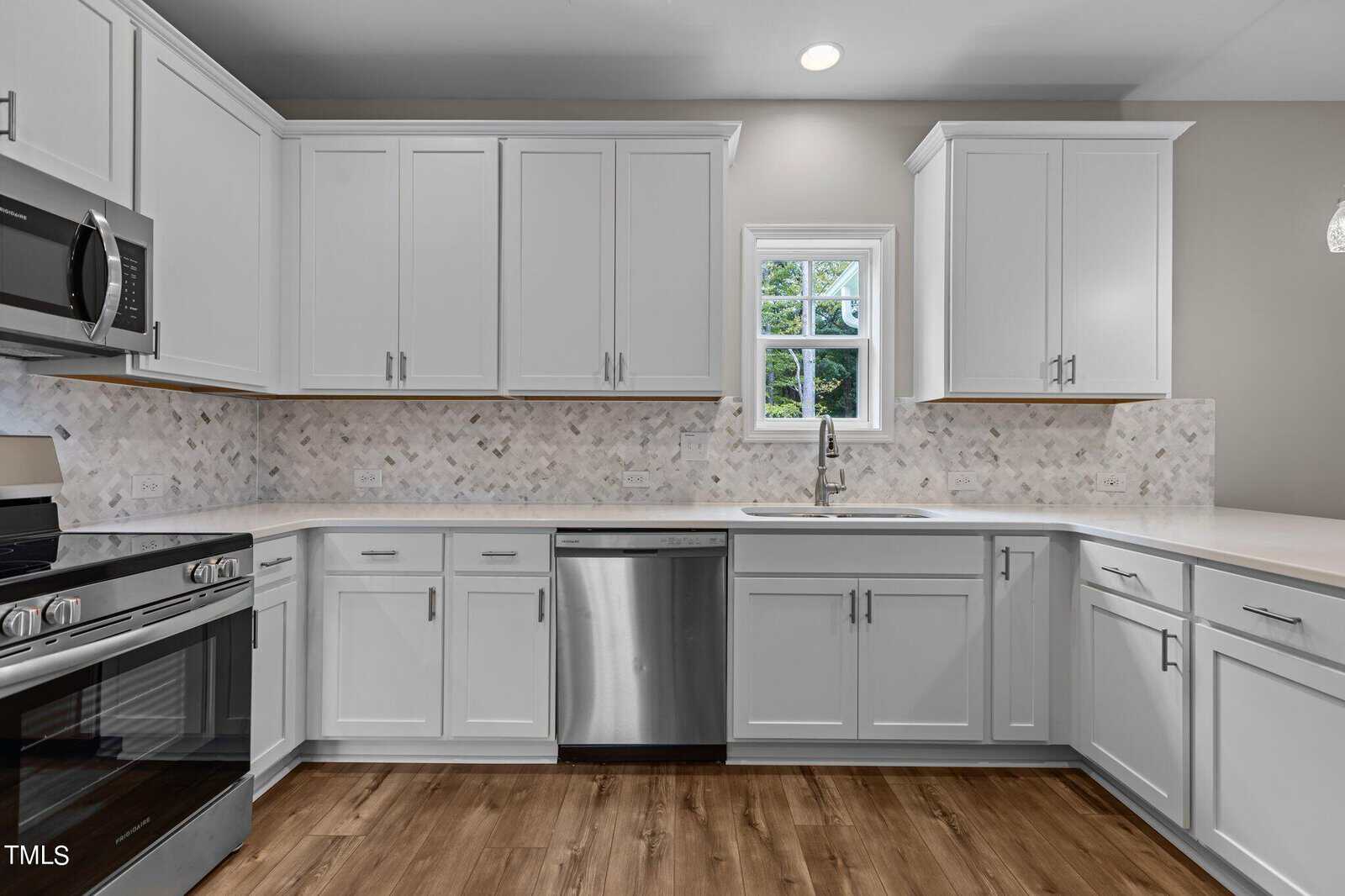 Modern kitchen with white cabinets, stainless steel appliances, a marble tile backsplash, a single window above the sink, and wood flooring. The space is bright and clean with neutral colors.