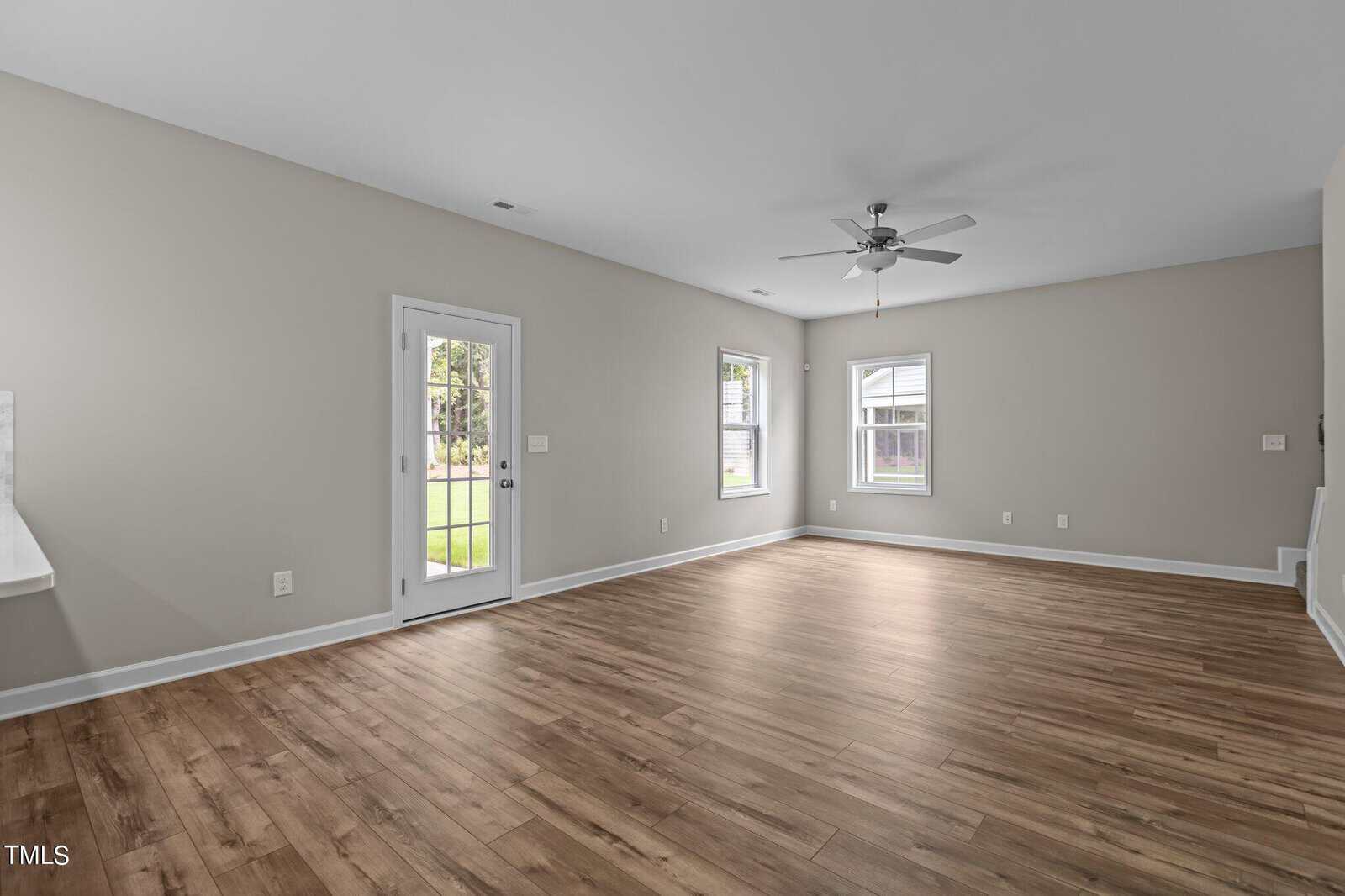 Spacious empty living room with light gray walls, wood flooring, ceiling fan, two windows, a glass door to the backyard, and white trim. Natural light fills the room, creating a bright and airy atmosphere.