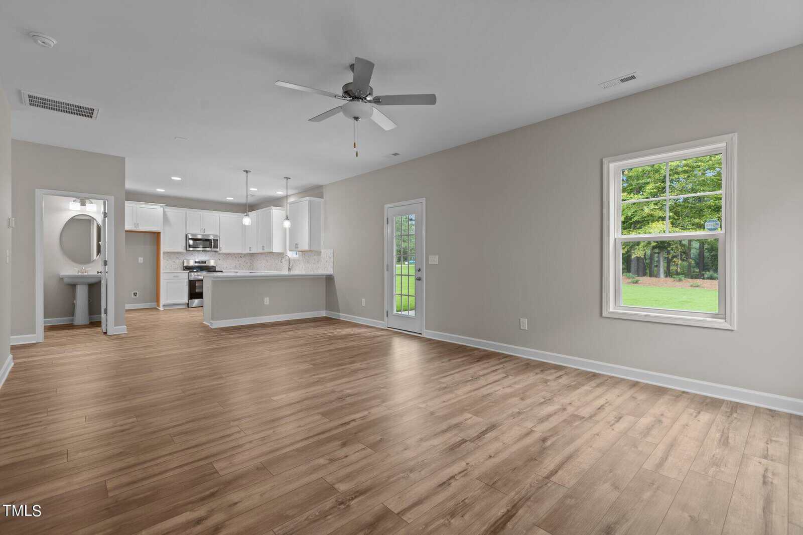 Open-concept living area with light wood flooring, neutral walls, ceiling fan, large window showing greenery, and a modern kitchen with white cabinets and stainless steel appliances in the background.
