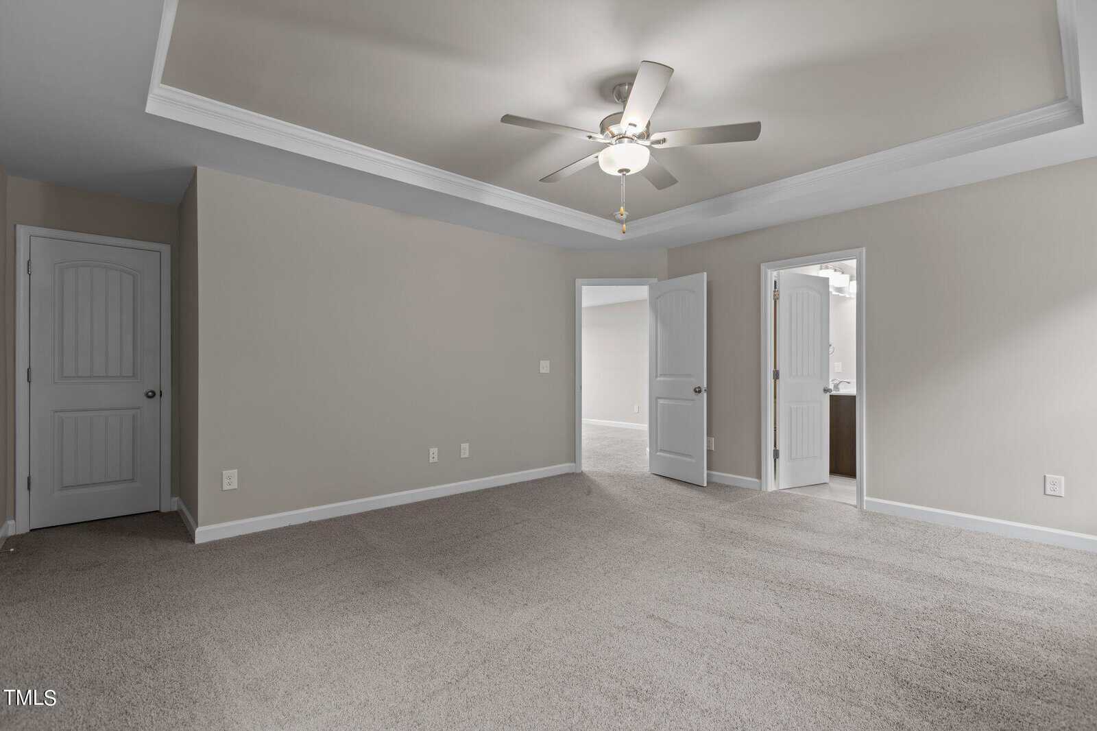 Spacious empty room with beige carpet, light gray walls, ceiling fan with light, and two open doors leading to other rooms. Crown molding accents the tray ceiling.