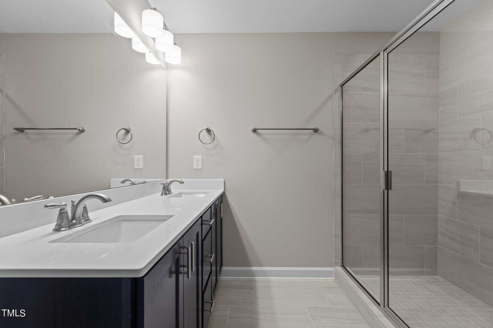 Modern bathroom with double sinks, large mirror, dark cabinets, and a glass-enclosed walk-in shower. The walls and floor are light-colored, giving the space a clean, minimalist look.