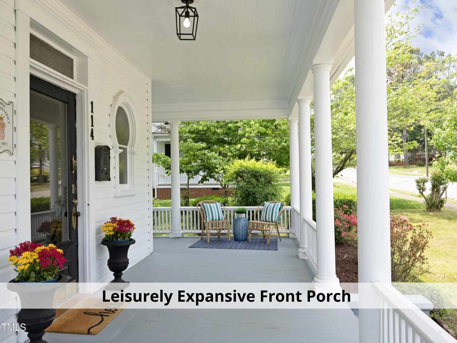 A spacious front porch with white pillars and a blue ceiling, featuring a cozy seating area with striped chairs and a small table. Potted flowers add color, and lush greenery surrounds the area. A mailbox and house number are visible.