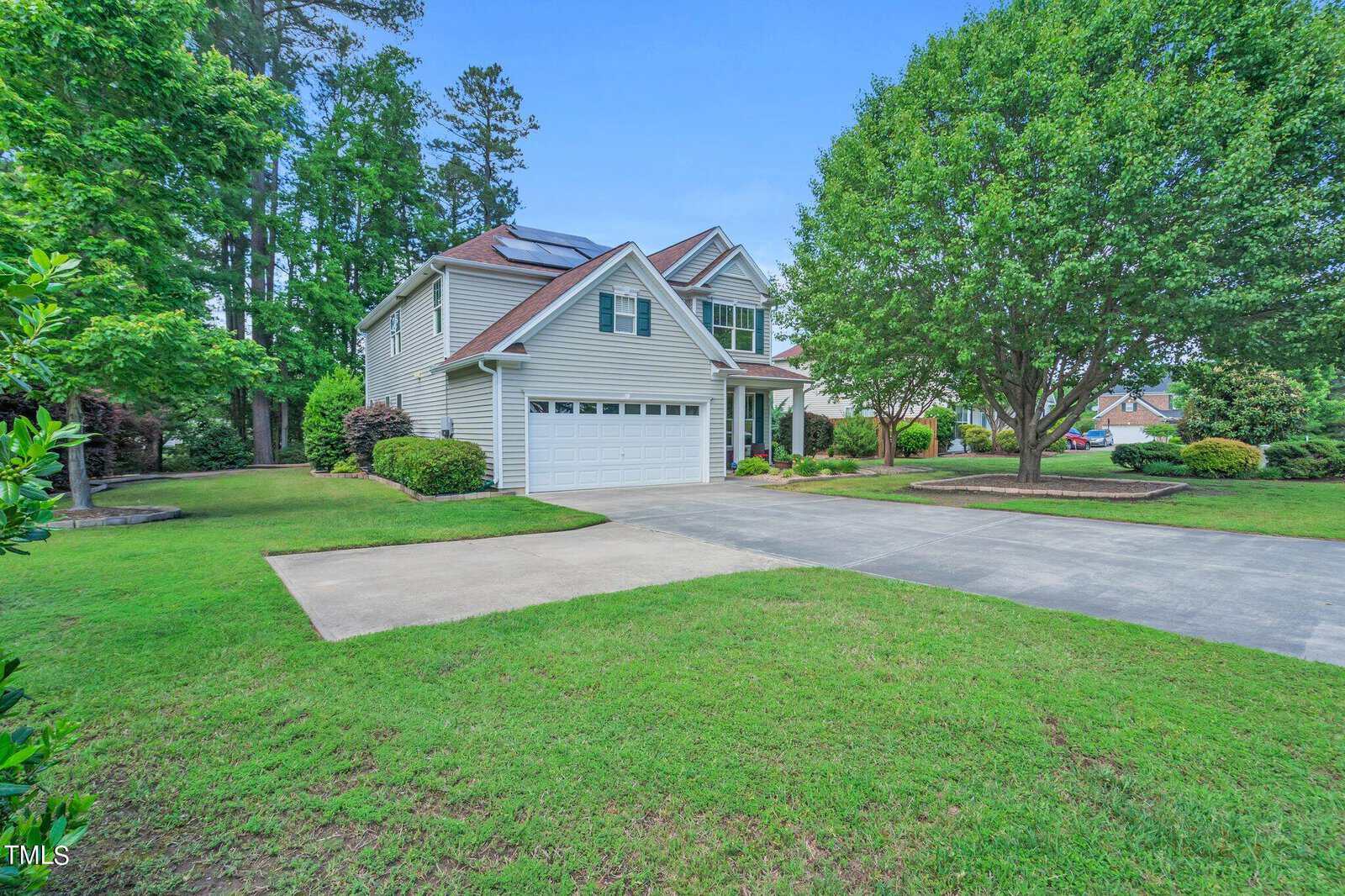 A two-story house with solar panels on the roof, a double garage, and a neatly manicured lawn. Trees and shrubs surround the property, and a driveway leads up to the garage. The sky is clear and blue.