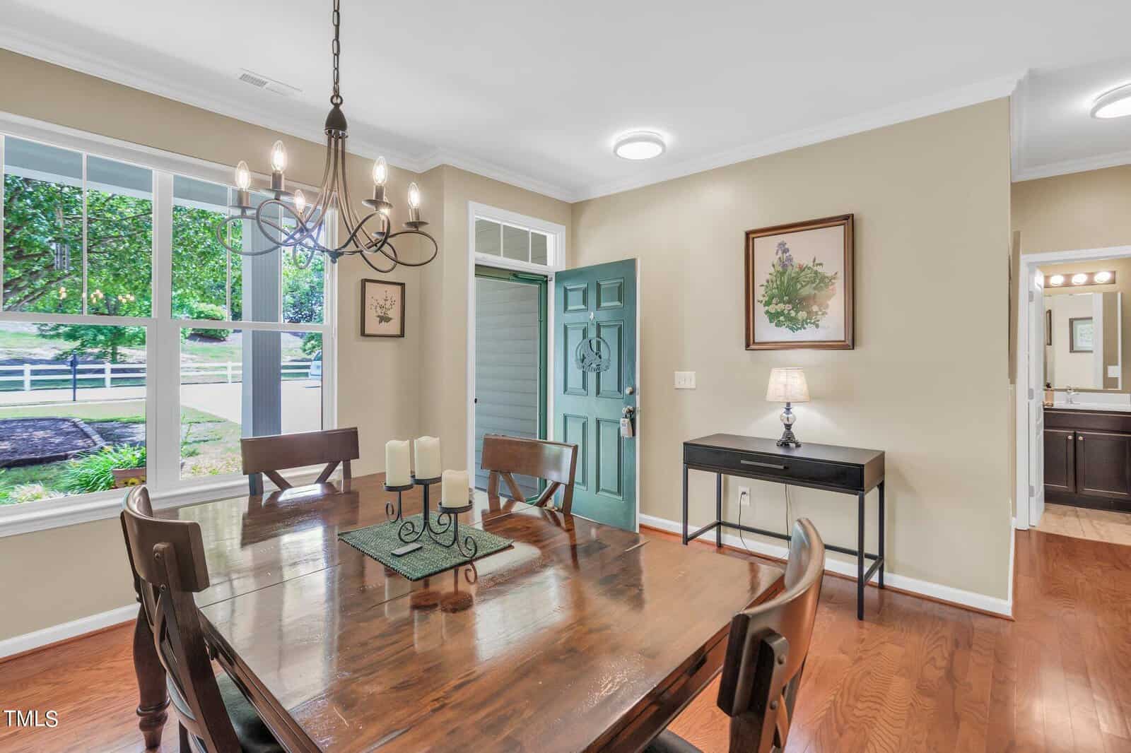 Dining room with wooden table and six chairs. A chandelier hangs overhead, and a table centerpiece with candles is present. The room has large windows, an open door, artwork on beige walls, and a side table with a lamp.