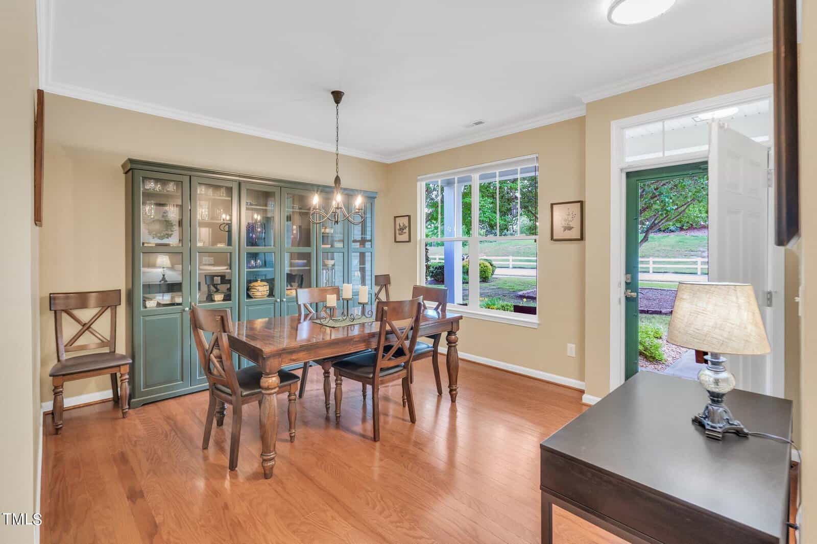 A dining room with a wooden table and six chairs, a green cabinet displaying dishes, and a candle centerpiece. Large window with a view of a fenced yard and trees. Hardwood floor, beige walls, white trim, and a lit chandelier.