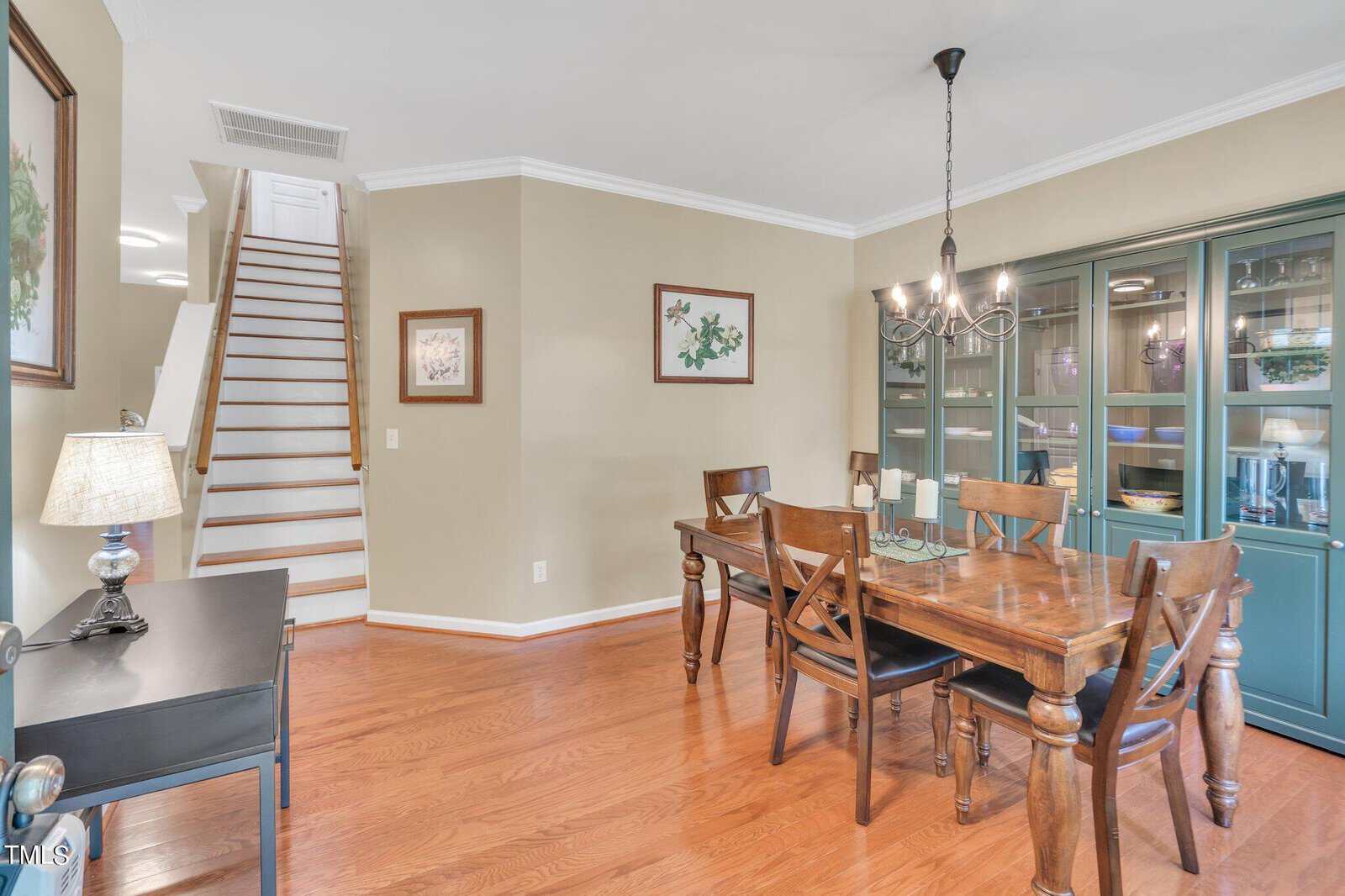Dining room with a wooden table and chairs, a black side table with a lamp, and a glass-front cabinet. The walls are adorned with floral artwork. Stairs with wooden steps and white risers are visible in the background. A chandelier hangs above the table.