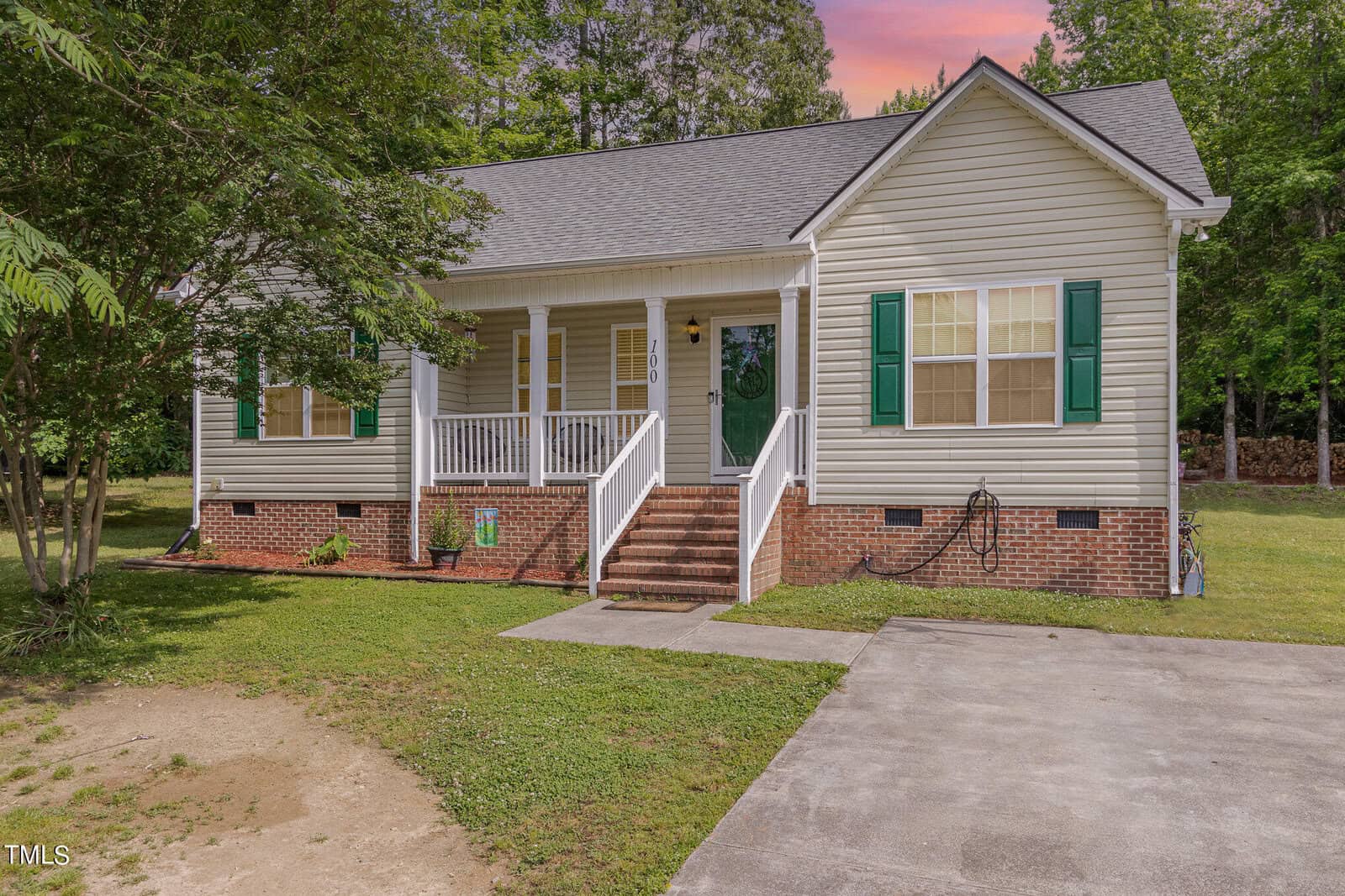 A single-story house at 100 Fir Court in Oxford boasts a brick foundation and beige siding, accented by green shutters and a dark green front door. The charming front porch with white railings overlooks a small grassy yard, while large trees frame the backdrop of a pinkish sky.