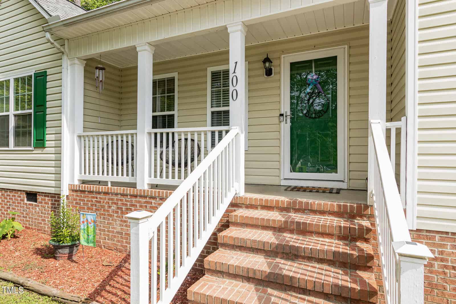A porch with white railings and brick steps leading to a green front door on a beige house. The home is surrounded by a small garden with plants and a brick-bordered flowerbed. House number "700" is displayed.