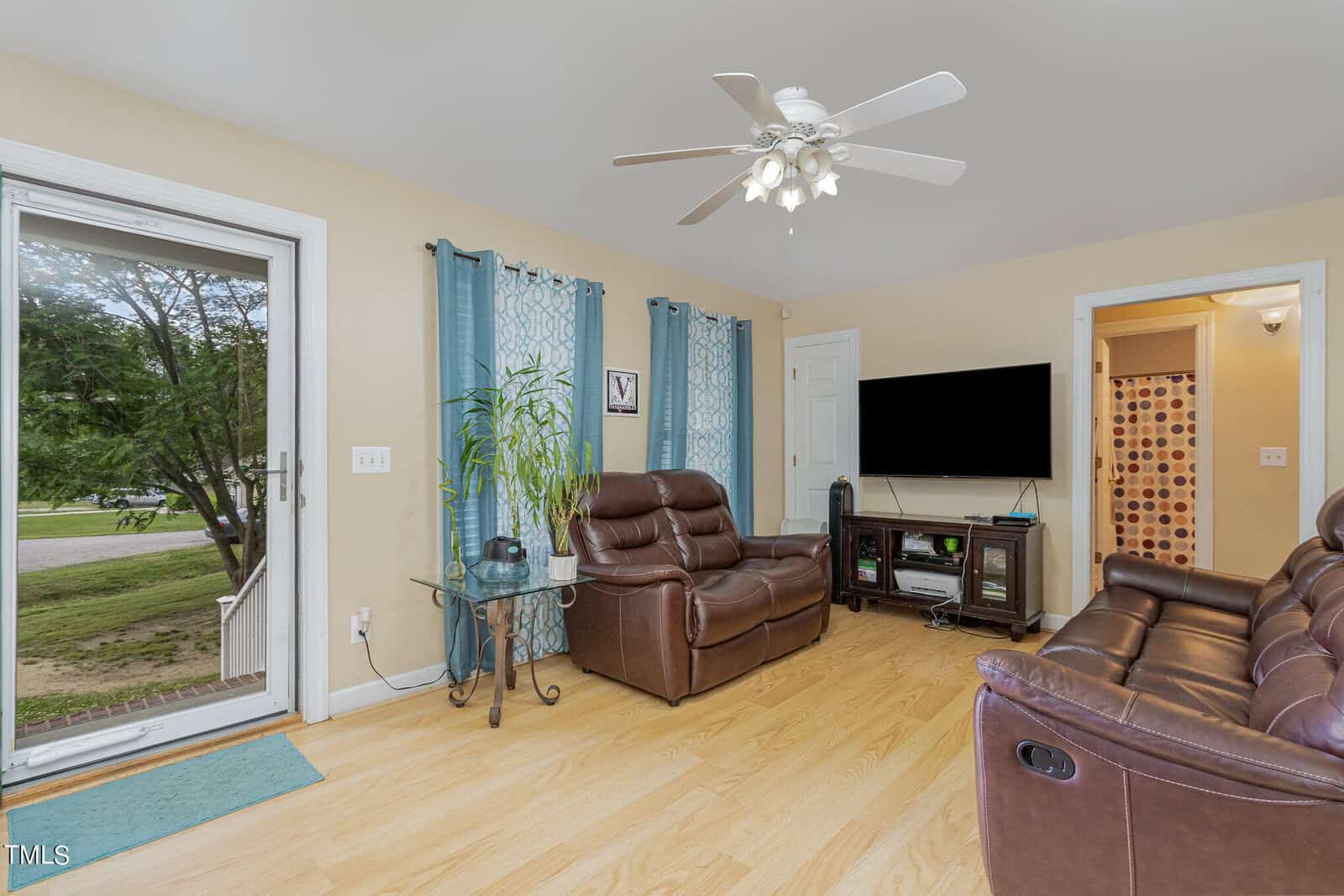 A cozy living room with brown leather sofas, a ceiling fan, and a flat-screen TV on a stand. There are blue curtains on two windows, a glass door leading outside, and a potted plant beside the sofas. The floor is light wood.
