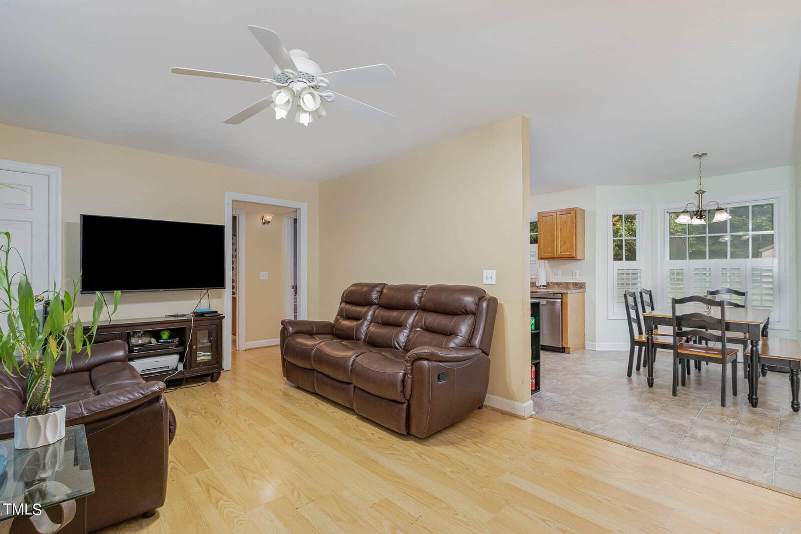 Spacious living room with brown leather sofas, a wall-mounted TV, and a ceiling fan. The room has wooden flooring and a few plants. An open doorway leads to a dining area with a table and chairs under a chandelier, and a view of the kitchen.