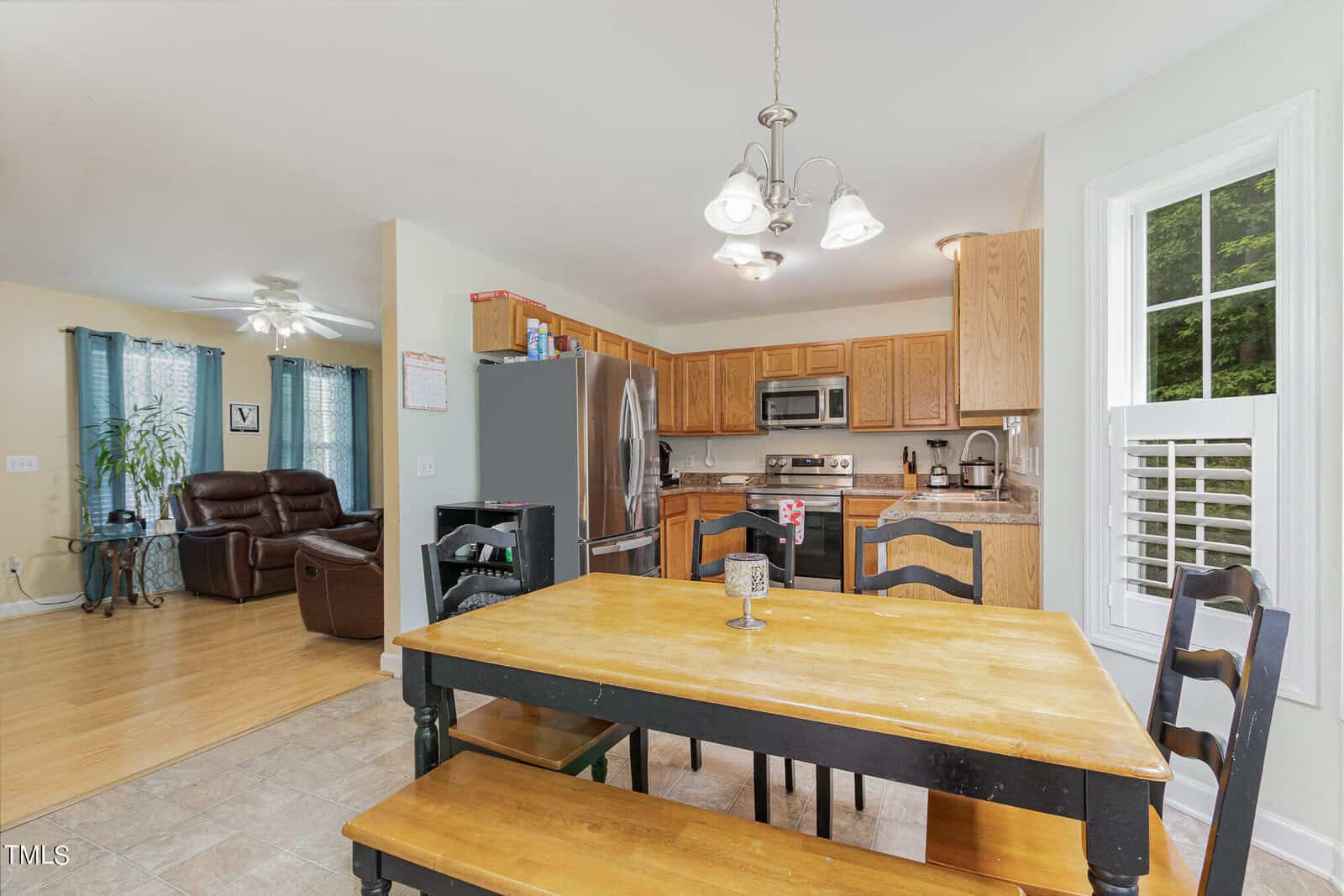 Bright kitchen and dining area with a wooden table and dark chairs. Stainless steel appliances and wooden cabinets in the background. Open layout leads to a living room with recliners and turquoise curtains. Light fixtures hang from the ceiling.