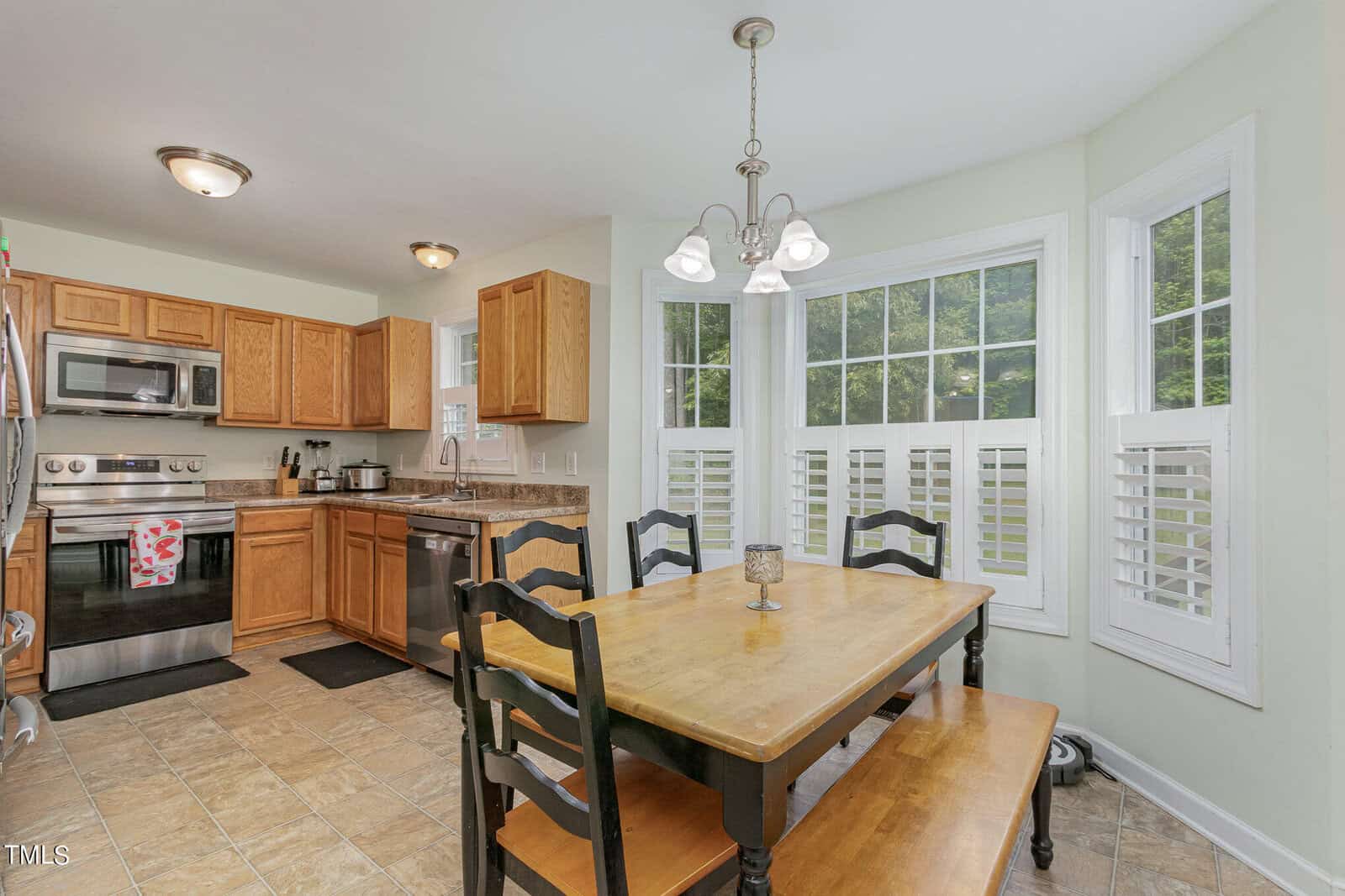 A kitchen and dining area with wooden cabinets and stainless steel appliances. A wooden table with black chairs and a bench is set in front of large windows. A chandelier hangs above the table, and light green walls complement the space.