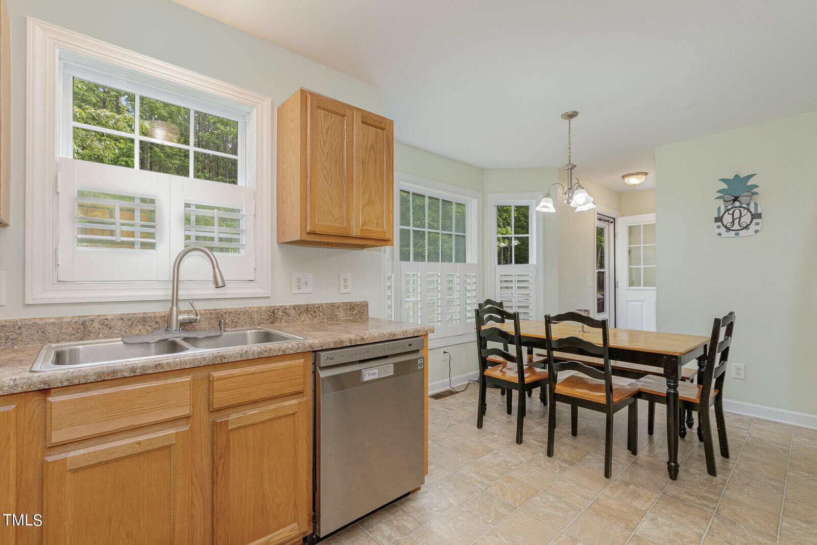 Bright kitchen with wooden cabinets, a stainless steel dishwasher, and a double sink under a large window. A dining area with a wooden table and four chairs is adjacent, lit by a small chandelier. The space has tiled flooring and white walls.