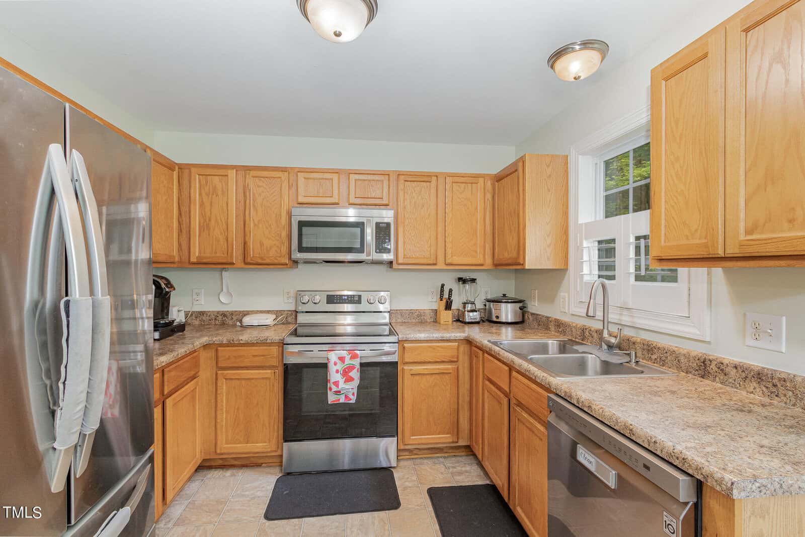 A modern kitchen with wooden cabinets, stainless steel appliances including a refrigerator, oven, microwave, and dishwasher. A double sink is below a window, and there are black floor mats. The countertop has various kitchen items.