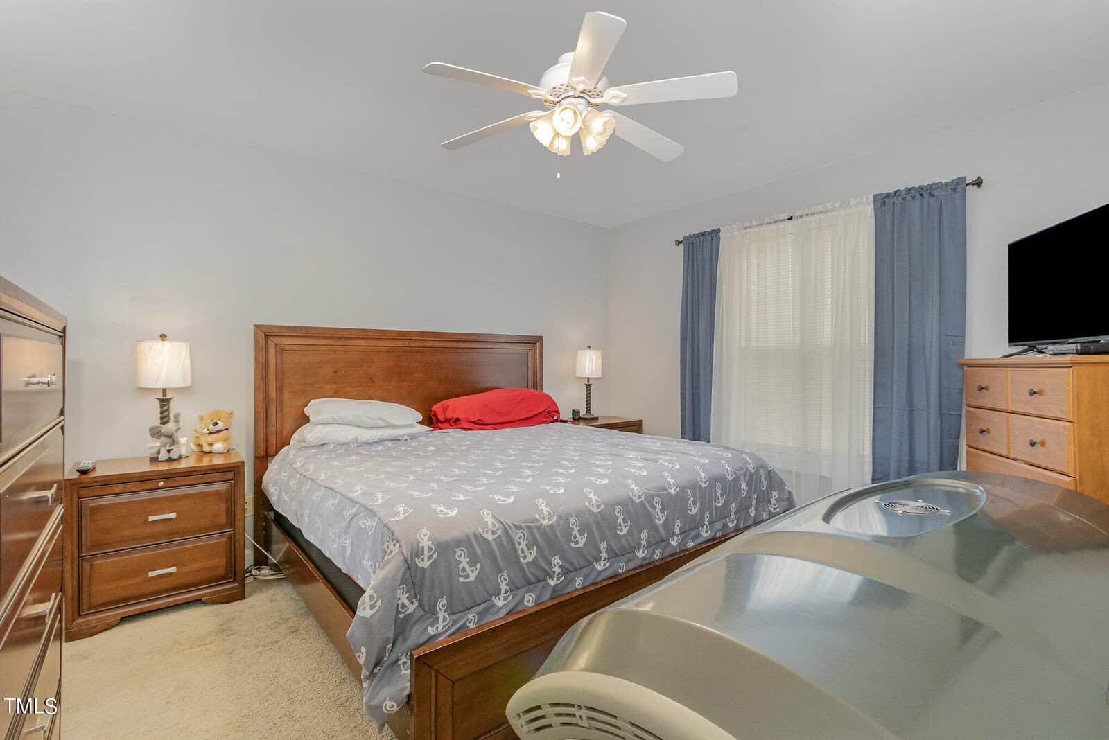 A bedroom with a wooden bed and blue anchor-patterned comforter, two bedside tables with lamps, a ceiling fan, a dresser with a TV, and blue and white curtains. A teddy bear and a red pillow add a cozy touch.