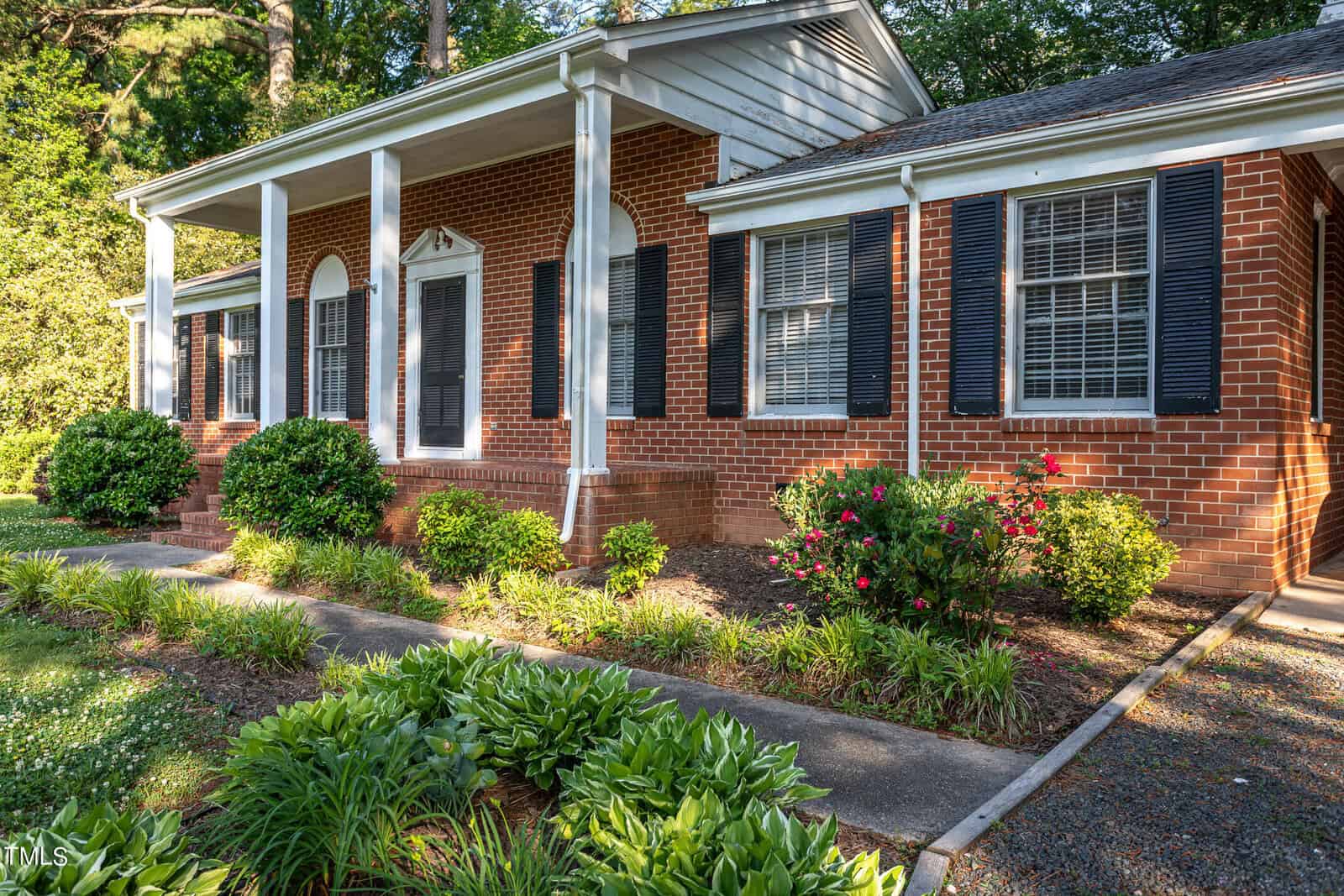 A traditional one-story brick house with white-trimmed windows and a covered porch, surrounded by lush greenery and flowering shrubs under dappled sunlight.