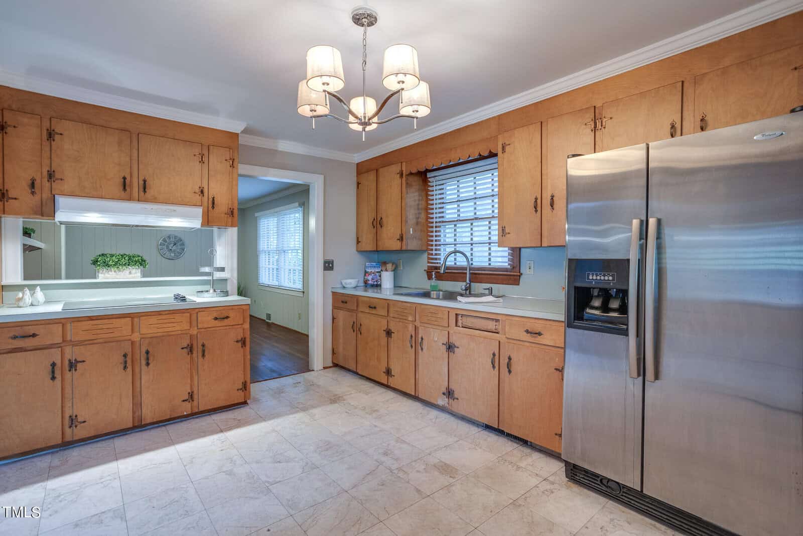 A spacious kitchen with wooden cabinets, a large refrigerator, and tile flooring. there’s a window with blinds above the sink and a serving hatch on the left.