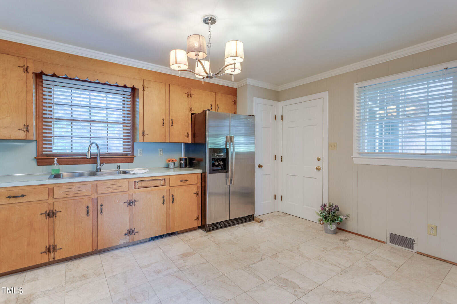 A spacious kitchen with wooden cabinetry, tile flooring, and modern appliances including a stainless steel refrigerator. there's a white door and window blinds, with a hanging light fixture.