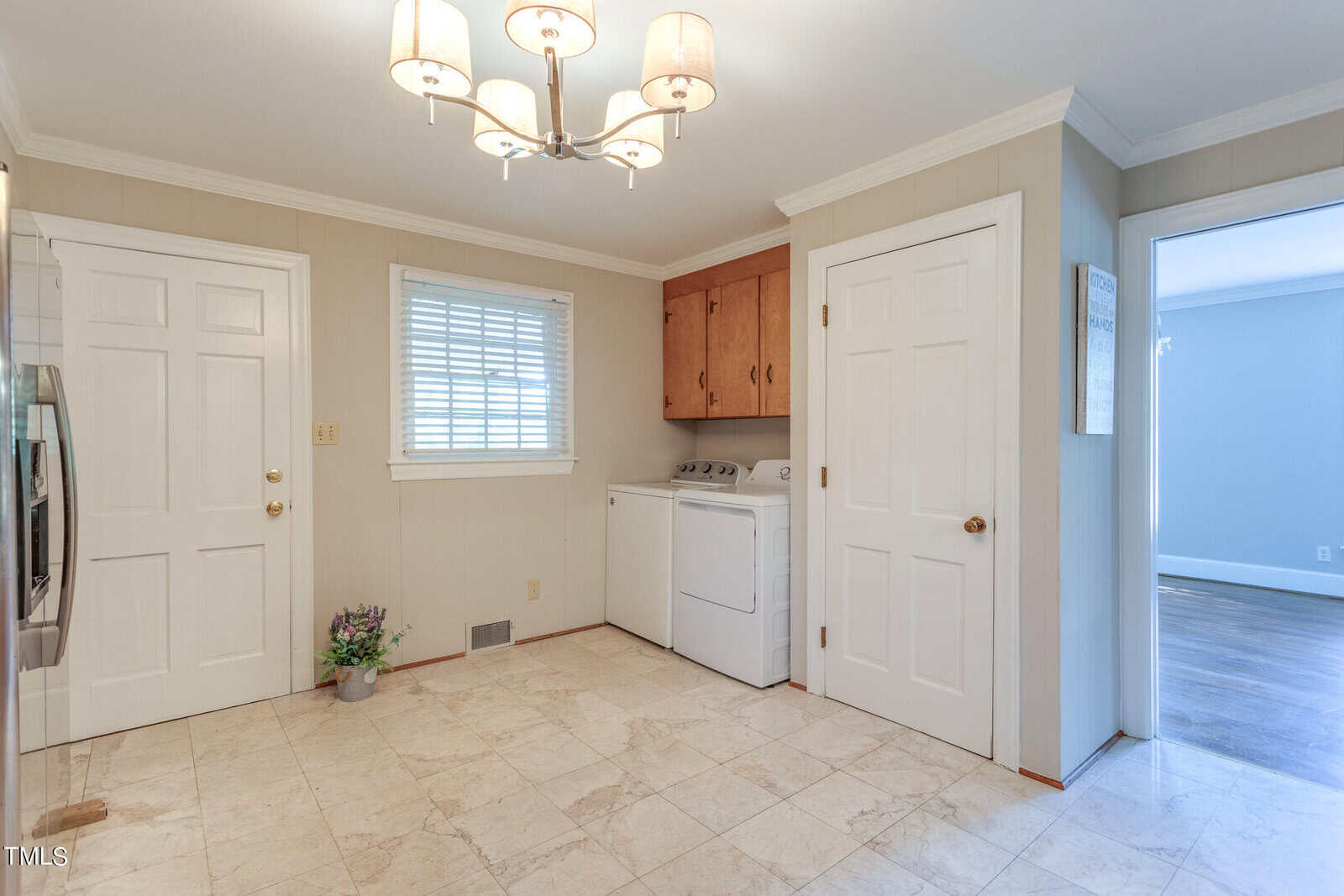A small, tidy laundry room with a washer, dryer, and wooden cabinet above. the room has beige floor tiles, a small window, and two white doors, one partially ajar. a pot of lavender sits on the floor.