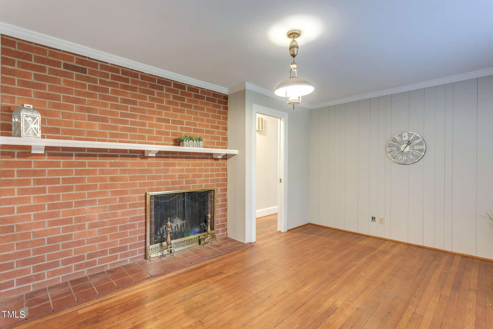 A bright room with a red brick fireplace, wooden flooring, and white-paneled walls. a mantle holds decorative items and a large silver clock hangs on the wall.