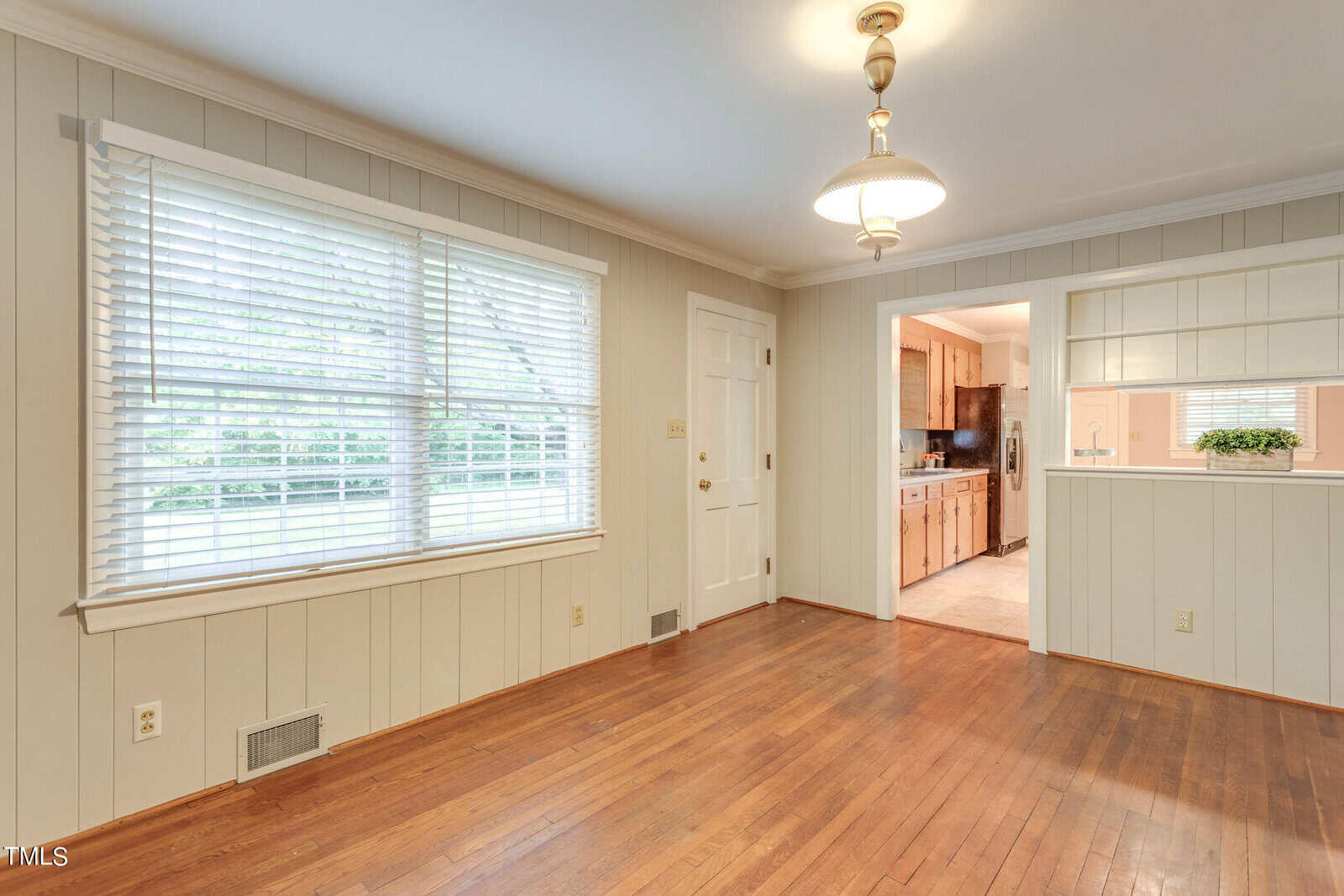 A bright and spacious room featuring a large window with white blinds, wainscoting on the walls, and hardwood flooring. a door leads to a kitchen visible in the background.