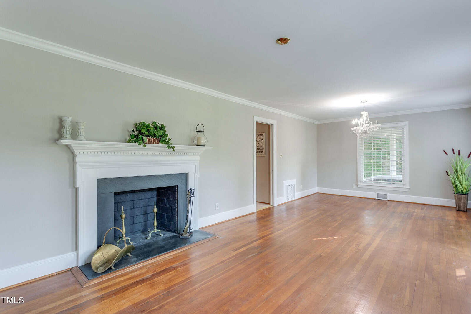 Spacious living room featuring a white fireplace with a mantel decorated with plants and a kettle, glossy hardwood floors, gray walls, and a large window with white trim.