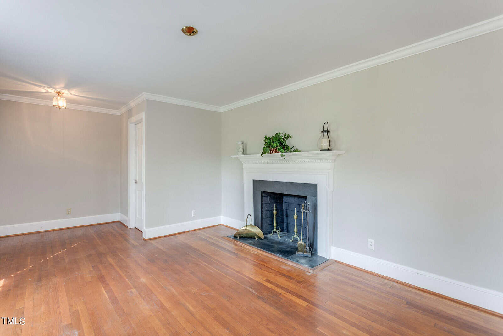 A spacious living room featuring hardwood floors, a white fireplace with a mantel decorated with a green plant, and light gray walls. neutral tones create a calm atmosphere.