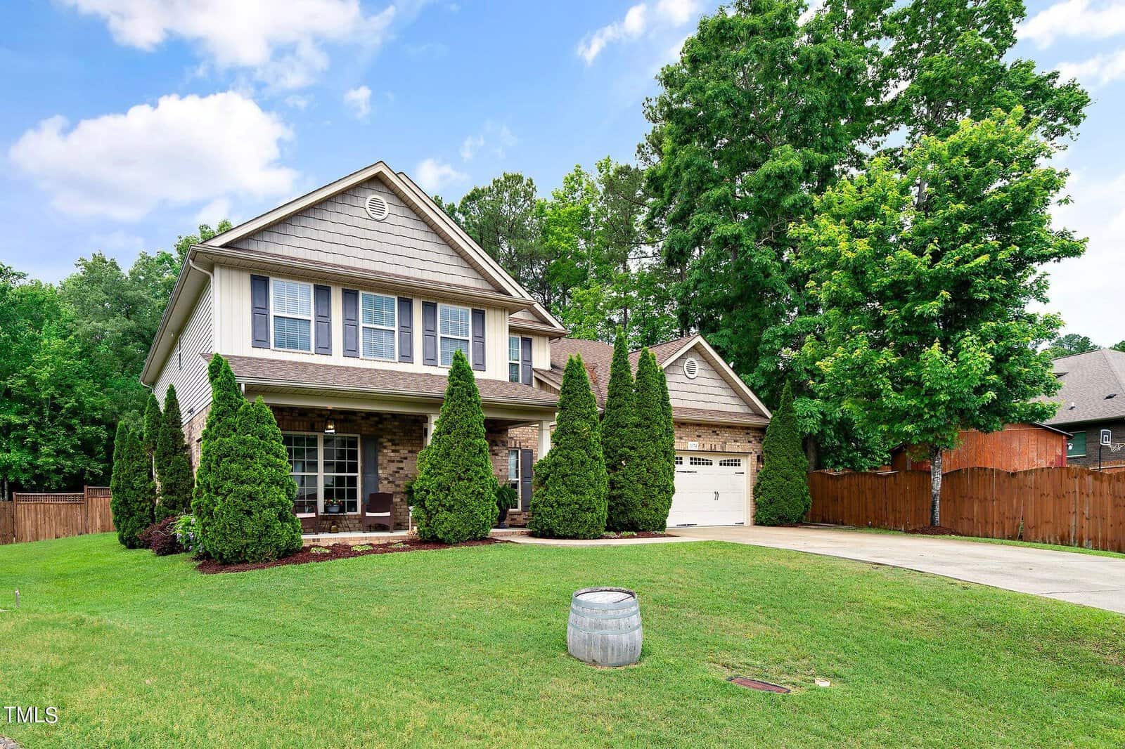 A two-story suburban house with a brick and siding exterior, surrounded by trimmed bushes and a manicured lawn. A wooden fence borders the property. The sky is clear with a few clouds.