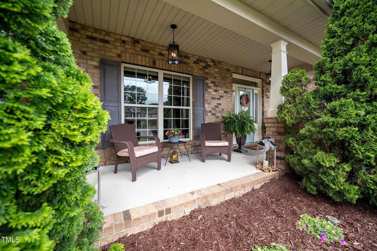 A cozy front porch with two wicker chairs and a small table on a light-colored surface. The house has brick walls, a large window, and a glass door adorned with a wreath. Green shrubs frame the porch, enhancing the inviting atmosphere.