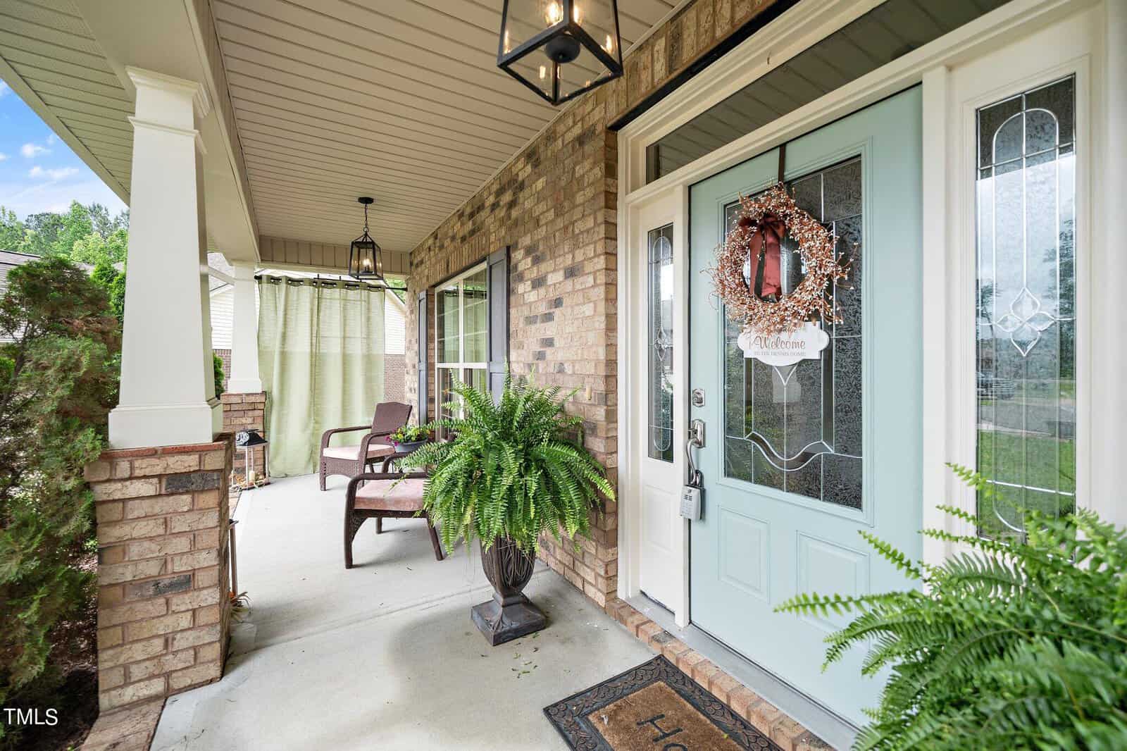 A welcoming porch with a light blue door adorned with a wreath and a sign. Brick walls and a white column frame the space. There are two brown chairs, a potted fern, and a patterned doormat that reads "Home." Hanging lights illuminate the area.