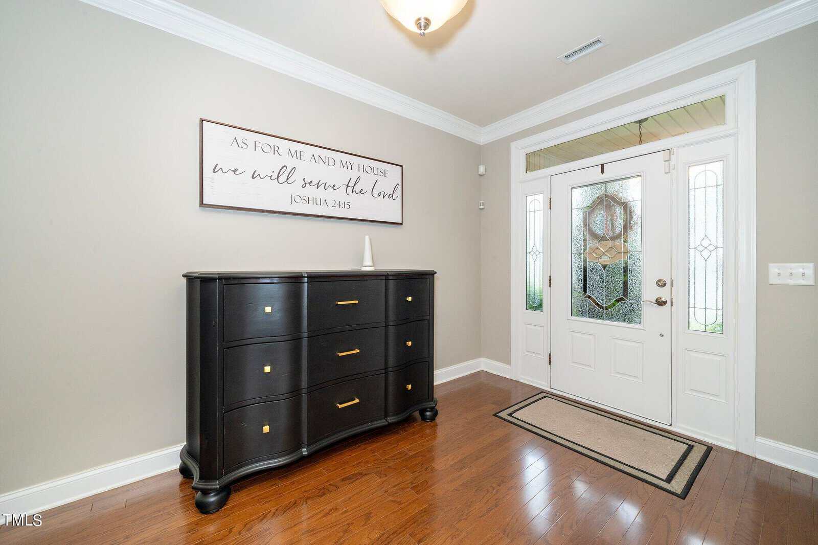 A bright entryway featuring a dark wooden dresser with gold handles on the left. A decorative sign with a biblical quote hangs above it. To the right, a white front door with glass panels and a wreath. The floor is polished wood.