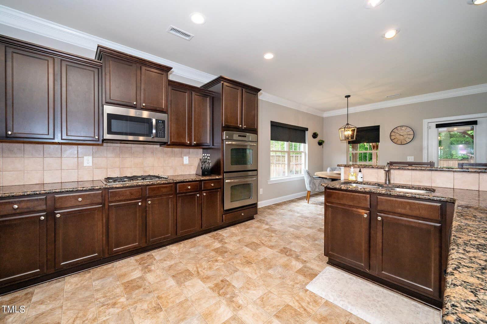 A modern kitchen with dark wooden cabinets, tiled floor, and granite countertops. Features include a built-in microwave, double oven, and stovetop. A breakfast bar separates the kitchen from a dining area with a pendant light and a window view.