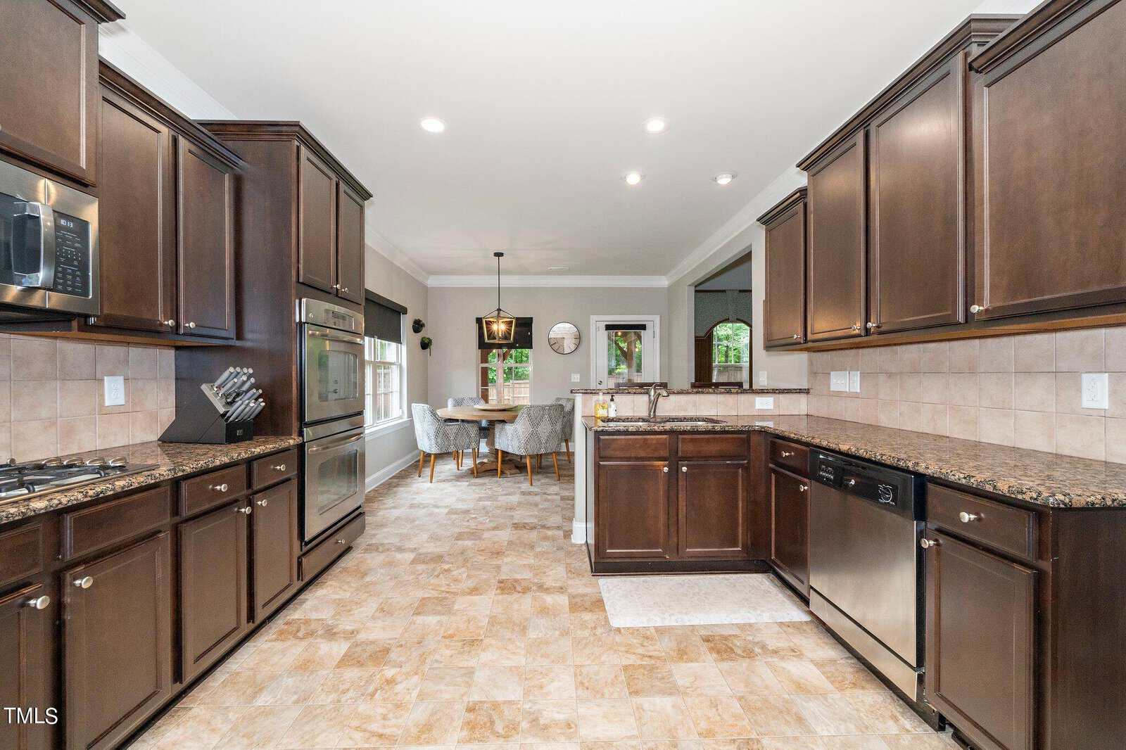 Spacious kitchen with dark wooden cabinets and granite countertops. Stainless steel appliances are visible, including an oven and dishwasher. There's a tiled floor and a dining area with a table and chairs by a large window in the background.