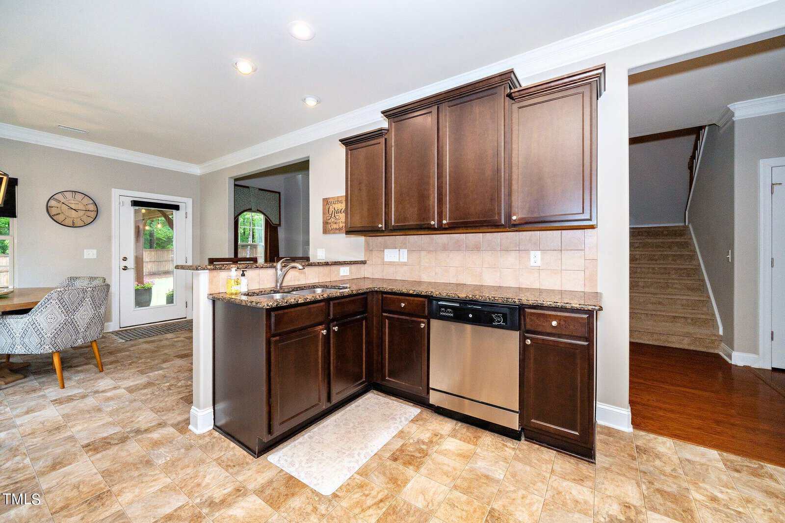 Spacious kitchen with dark wood cabinets, stainless steel dishwasher, and a tile backsplash. There's a sink on the island, beige tile flooring, and a view of a dining area with a large clock on the wall. Stairs lead to an upper level in the background.